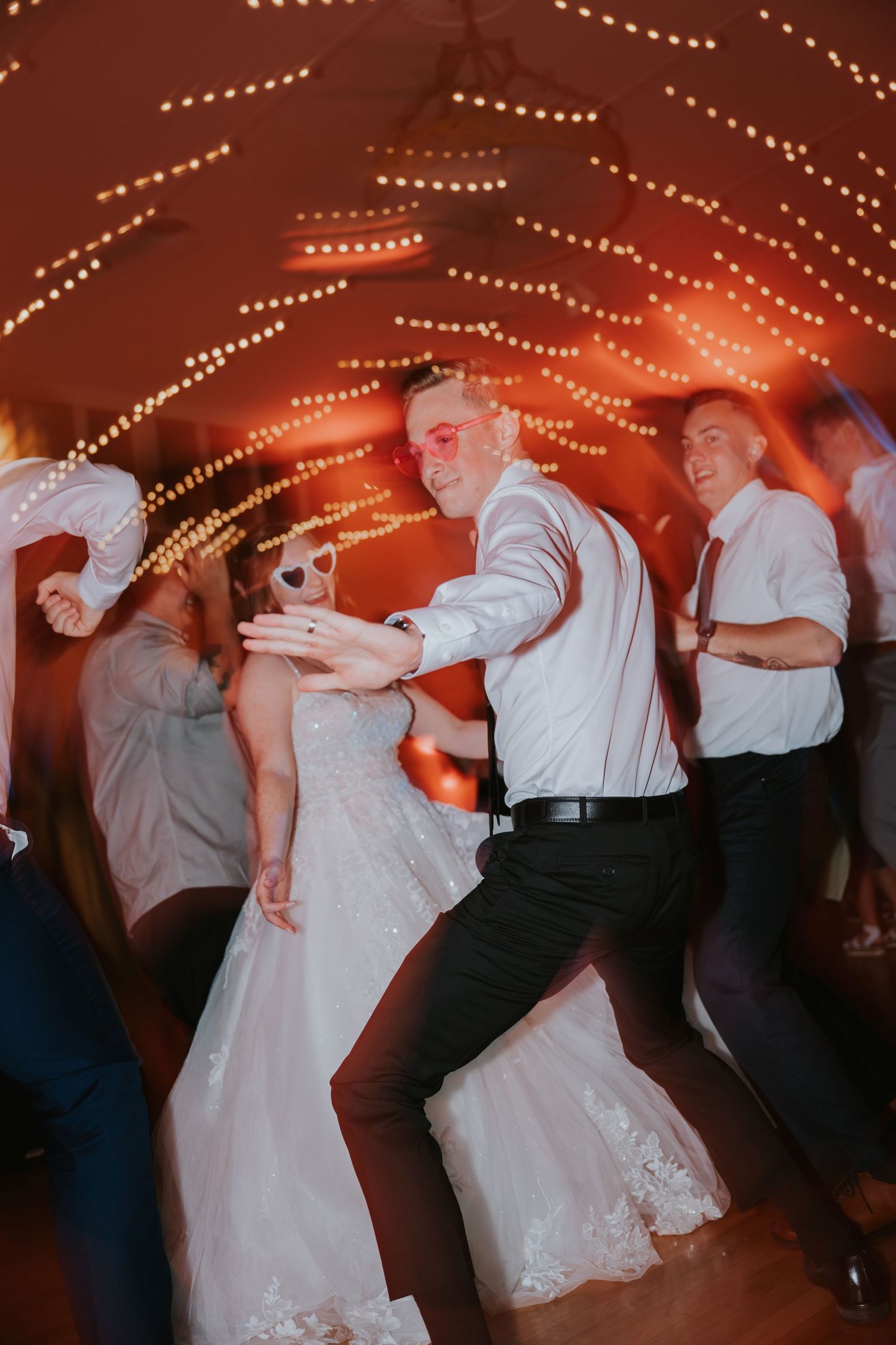Wedding guests dance together under warm string lights at a reception, creating a lively and joyful atmosphere.