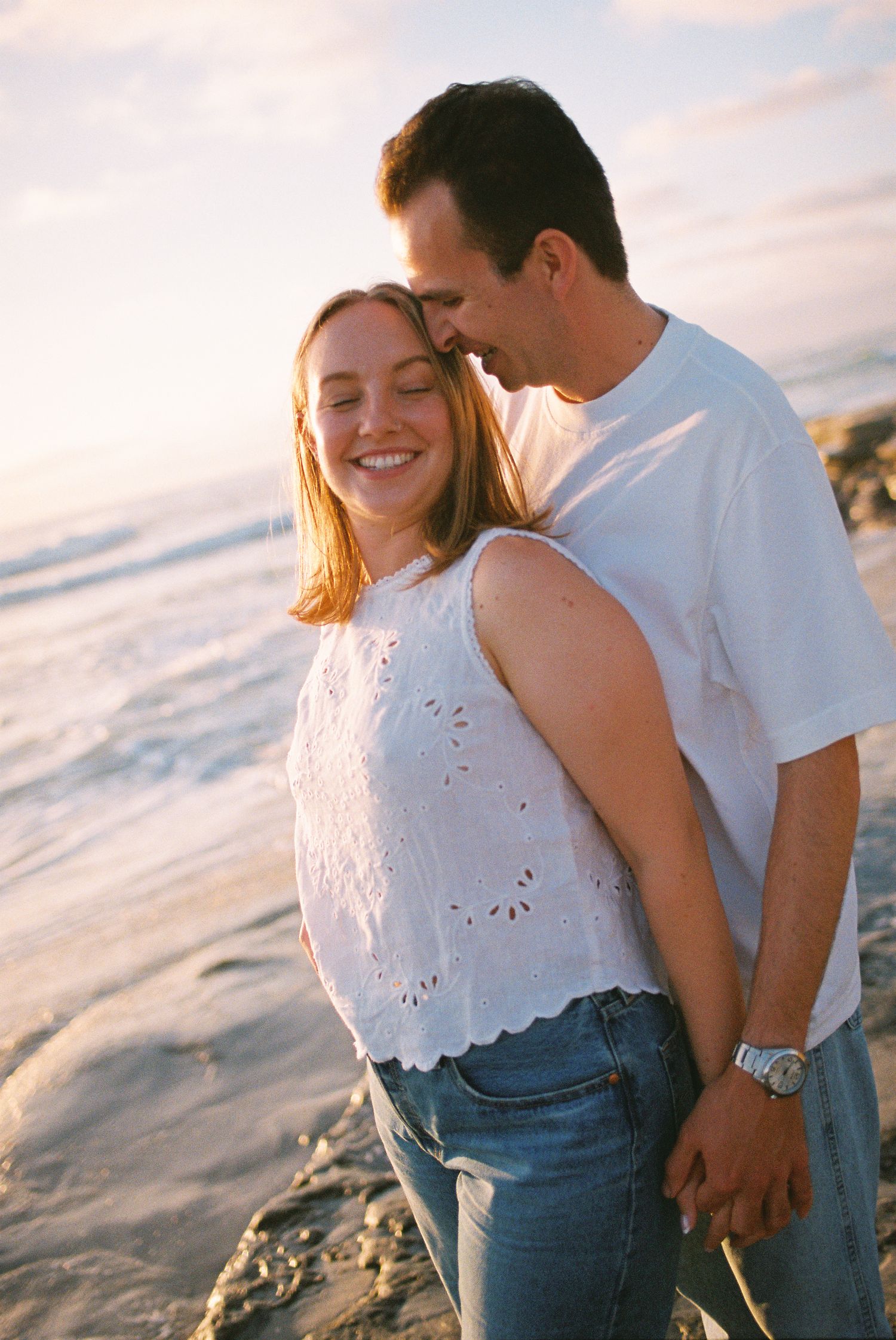 Happy couple in white tops and jeans embracing on a sunny beach, smiling together near ocean waves at golden hour.