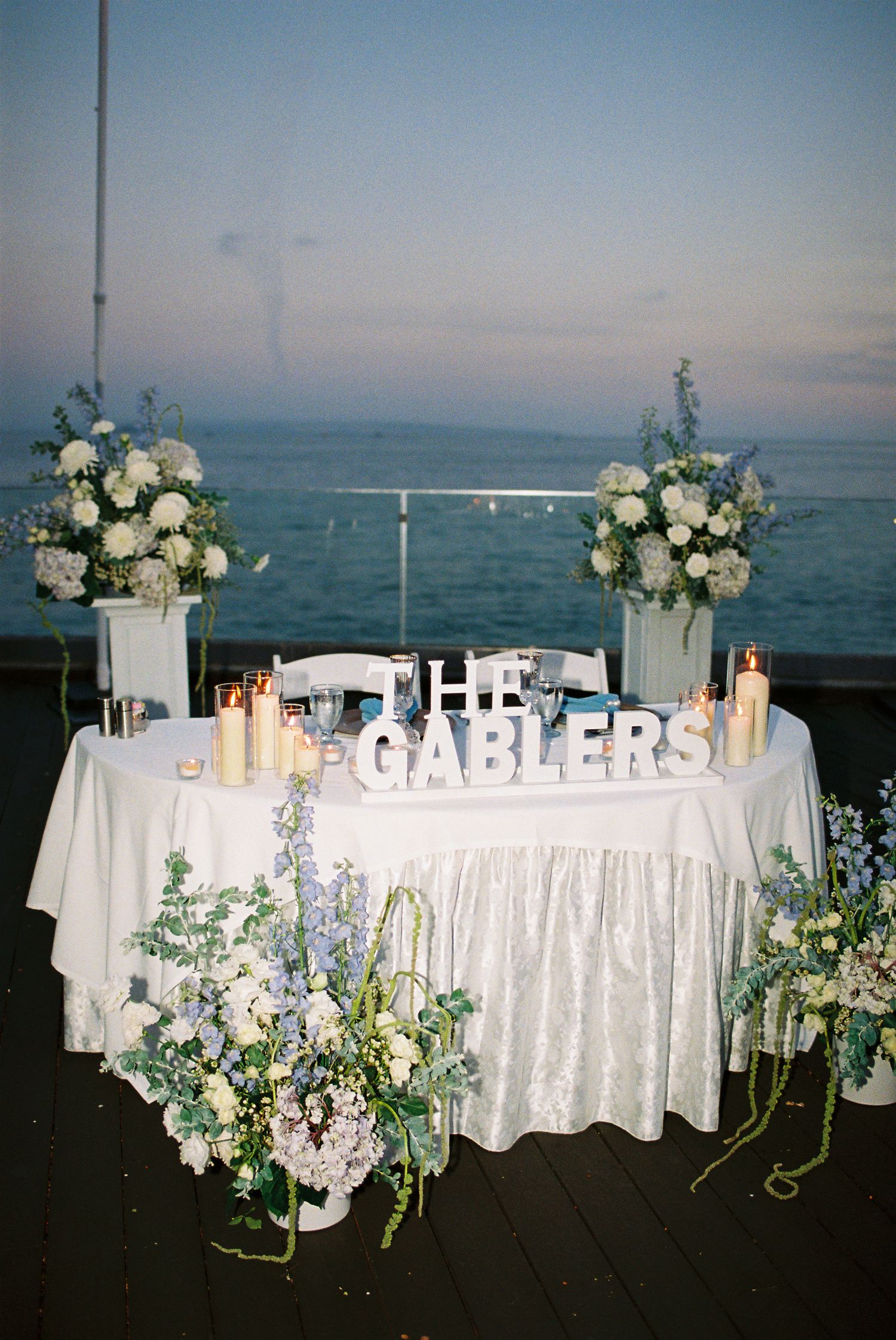 Elegant wedding sweetheart table for The Gablers with white floral arrangements and candles overlooking the ocean at dusk.