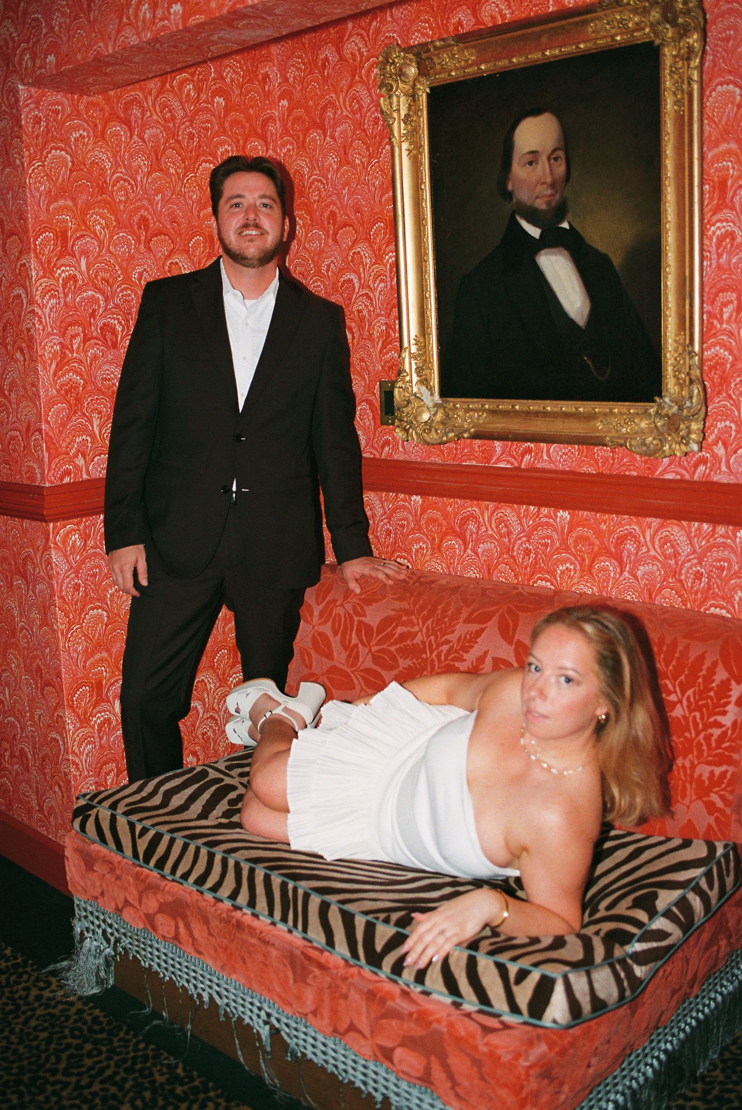 Man in black suit standing behind woman in white dress lounging on zebra print chaise beneath vintage portrait painting.