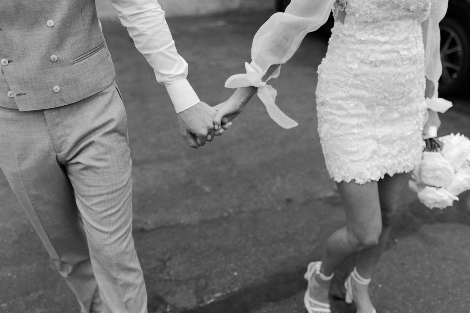A bride and groom walk hand in hand outdoors while showing off their wedding rings in a black and white photo.