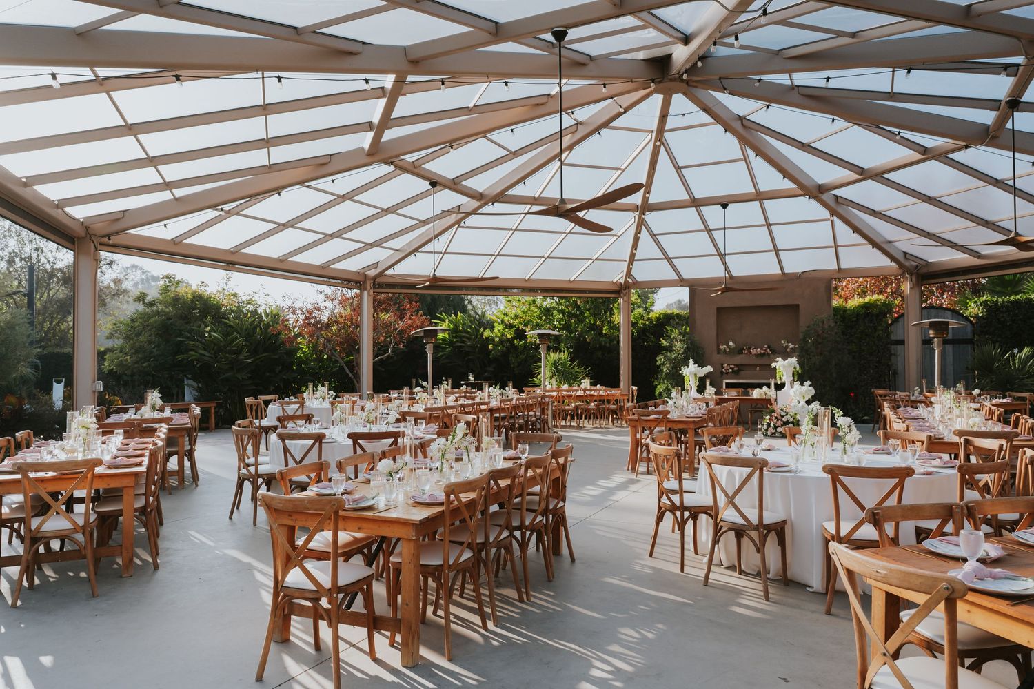 A panoramic view of an empty restaurant patio with wooden tables and chairs under a transparent roof covering.