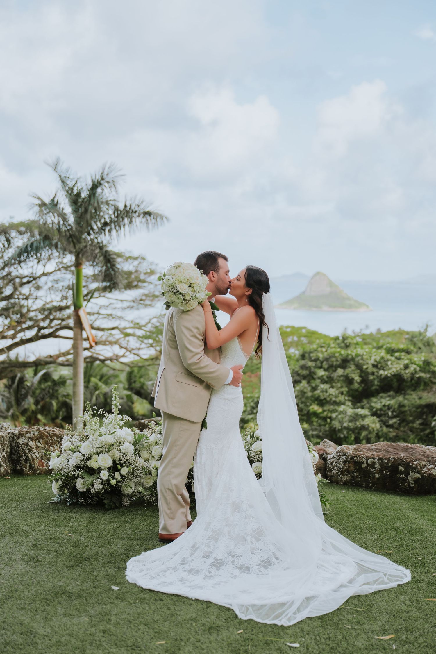 A romantic wedding moment captured on a scenic Hawaiian lawn with palm trees and mountains in the distance.