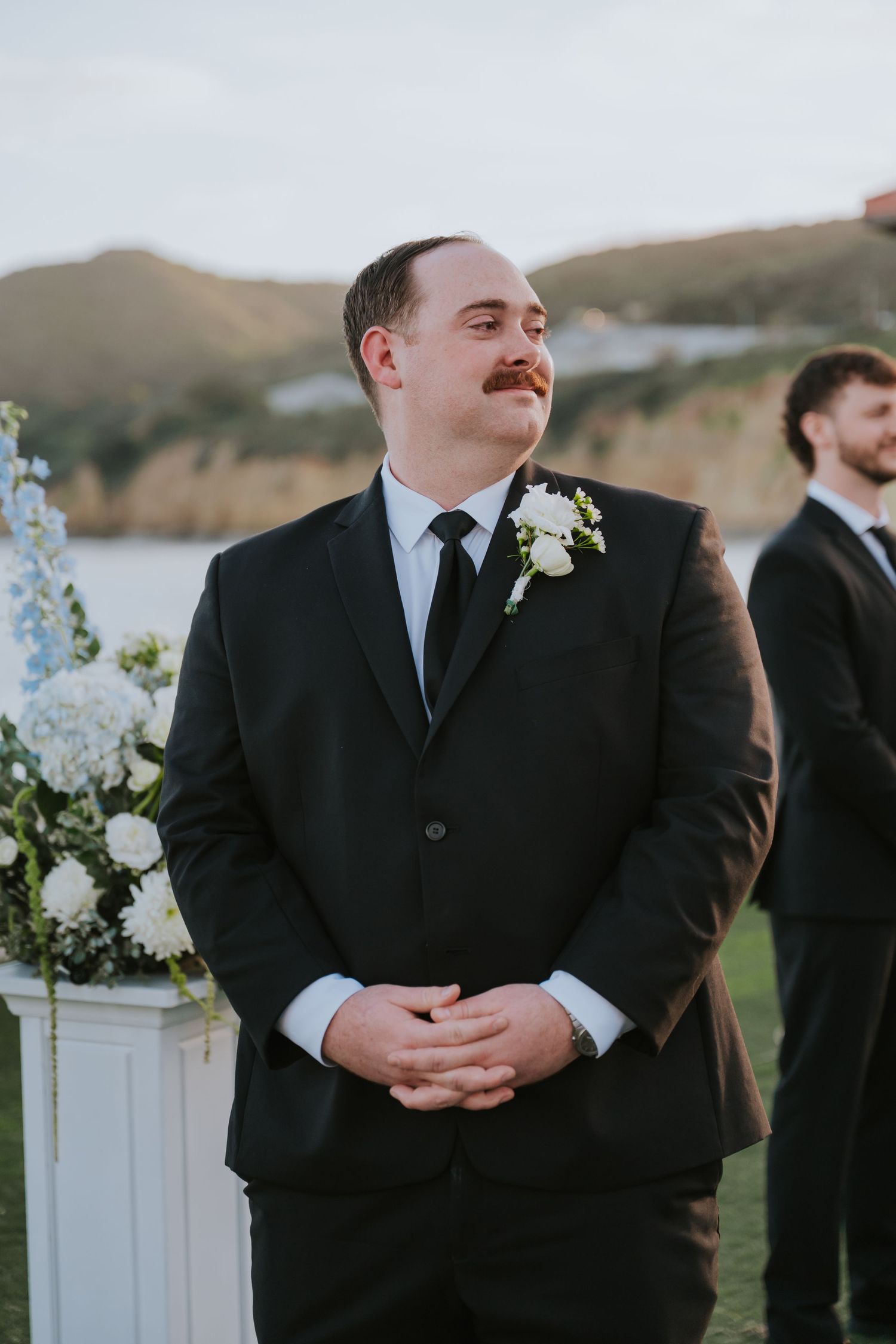 Groom in black suit with white boutonniere smiling at outdoor wedding ceremony with ocean backdrop.
