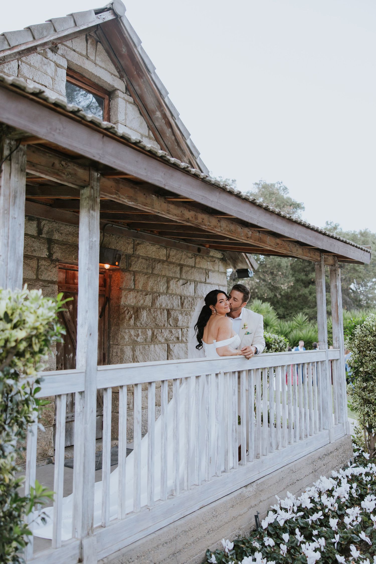 A bride and groom share a romantic moment on a rustic stone cottage porch surrounded by white flowers.