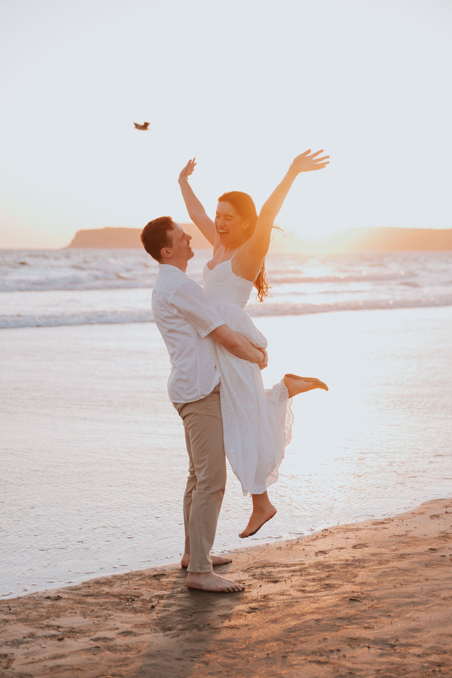 Joyful couple at beach sunset, man lifting woman in white dress with arms raised in celebration.
