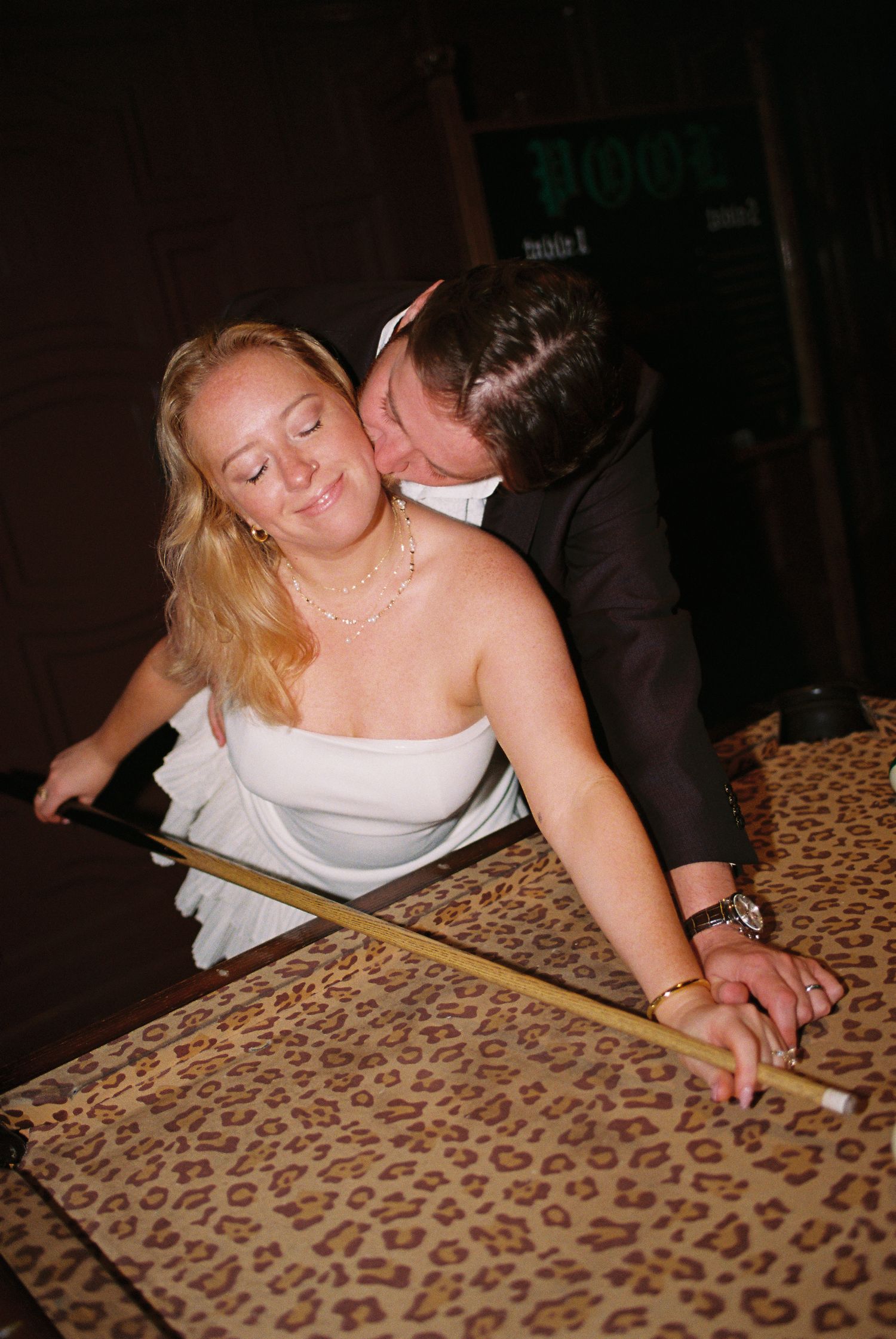 Bride in white strapless dress receives a kiss from groom in dark suit while leaning over a leopard print pool table.