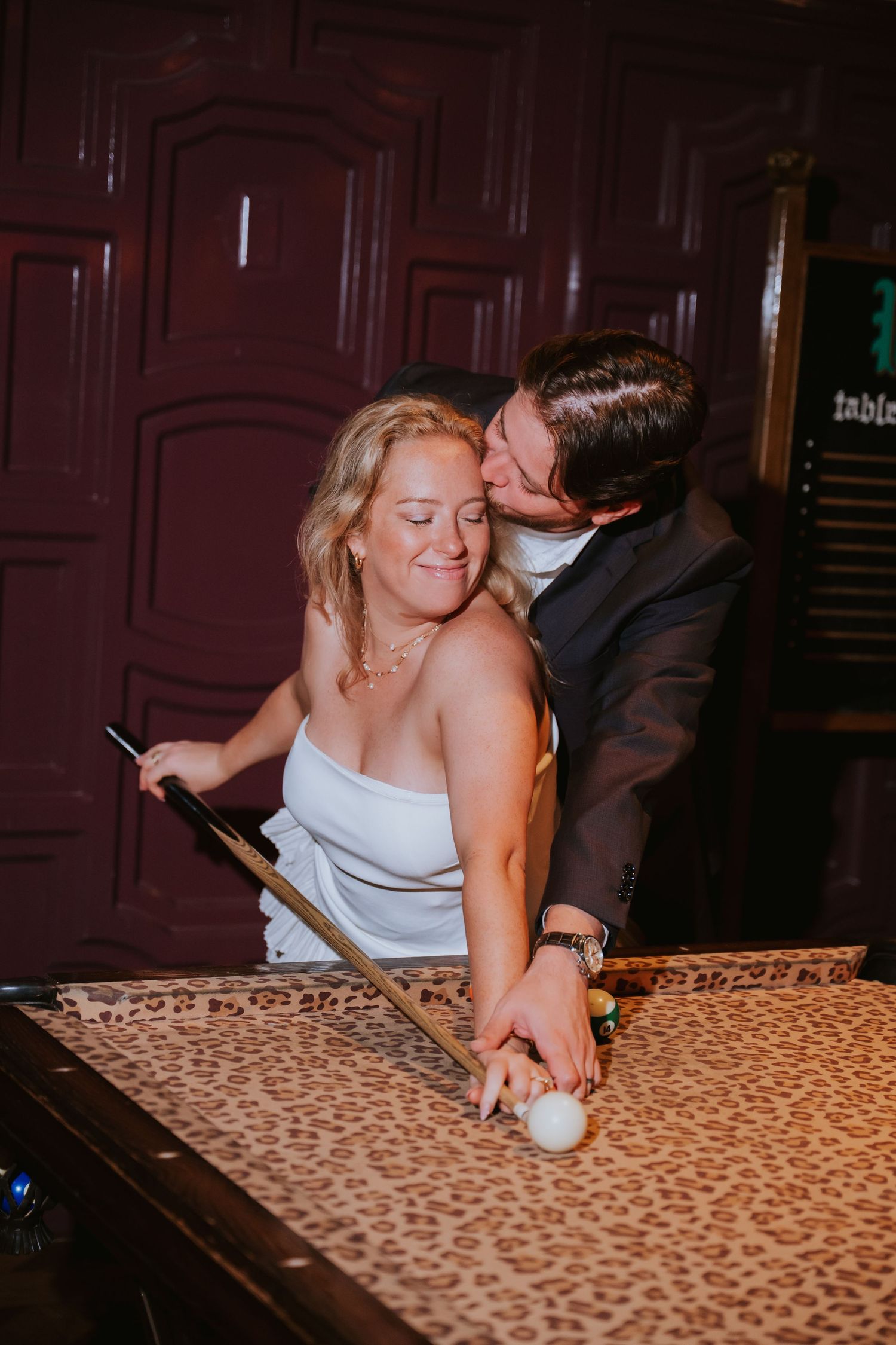 Groom kisses bride on cheek as she plays pool at leopard print billiard table during wedding reception.