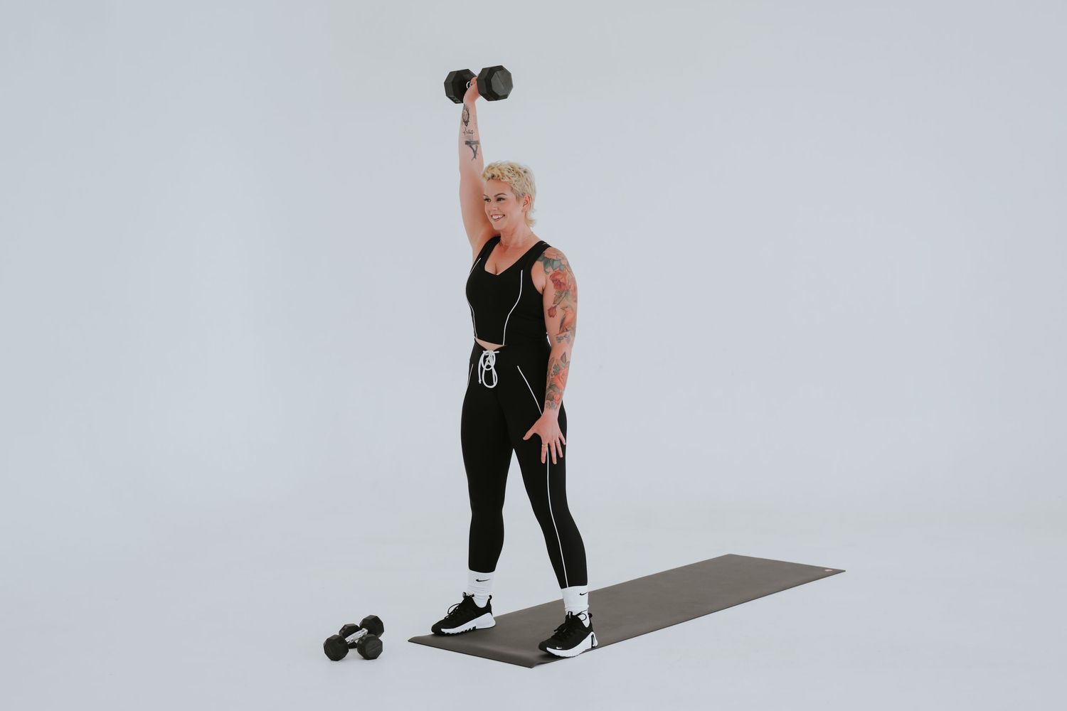 Person in black workout attire performs standing dumbbell exercise on yoga mat against white background.