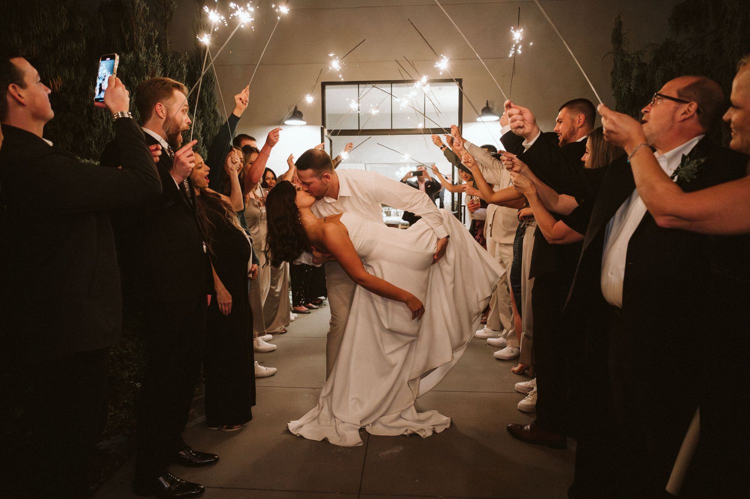 Wedding couple shares a romantic kiss during sparkler exit while guests hold glowing sparklers in a dark venue.