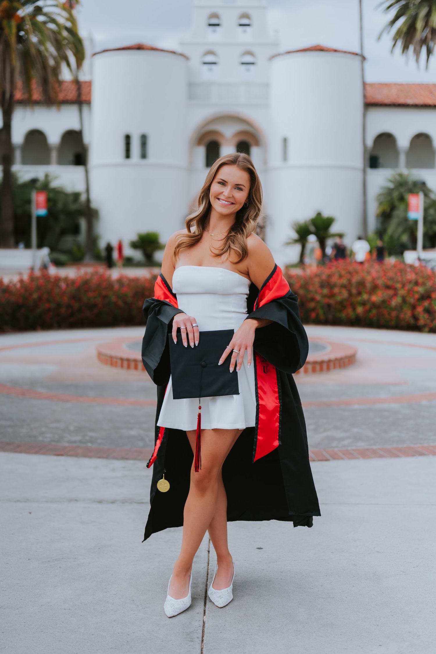 Young woman in white dress and graduation gown holds diploma outside a Spanish-style university building.