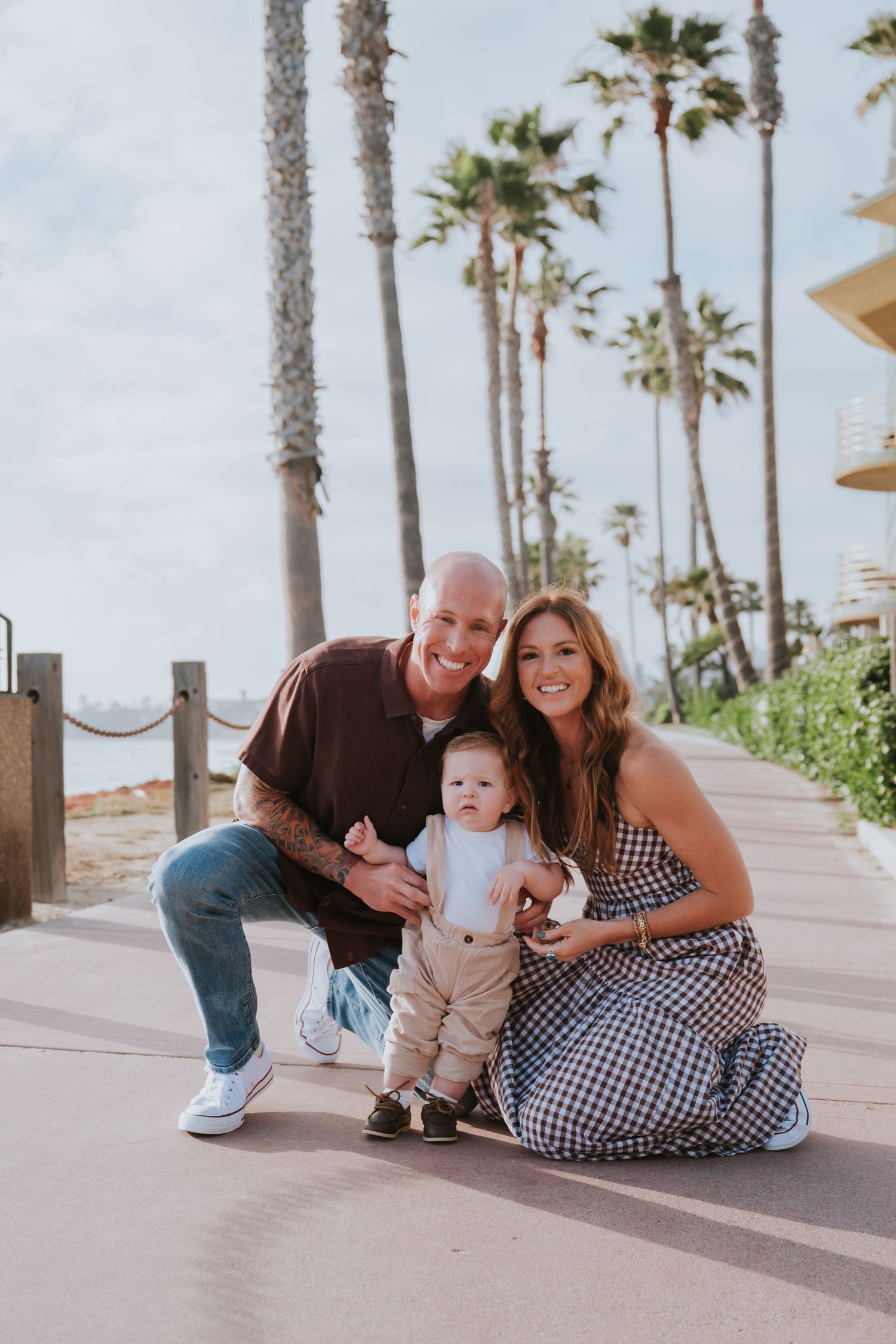 Young family with baby posing on sunny beachside path with tall palm trees in background.