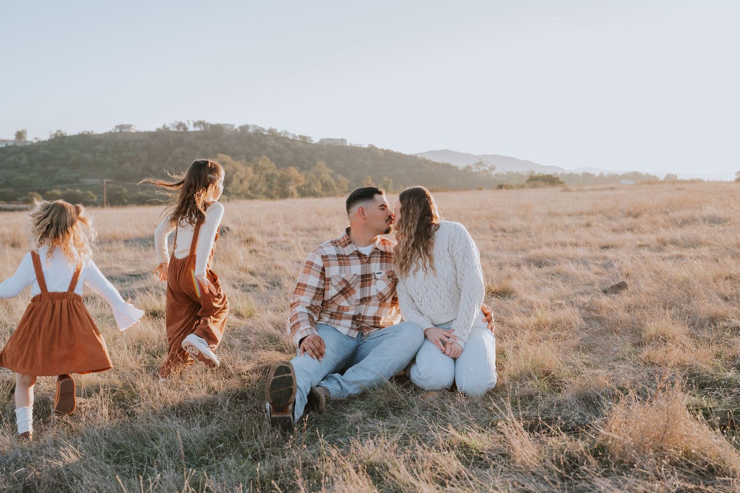 A couple shares a kiss in a golden field while two young girls in rustic dresses play nearby under warm sunlight.