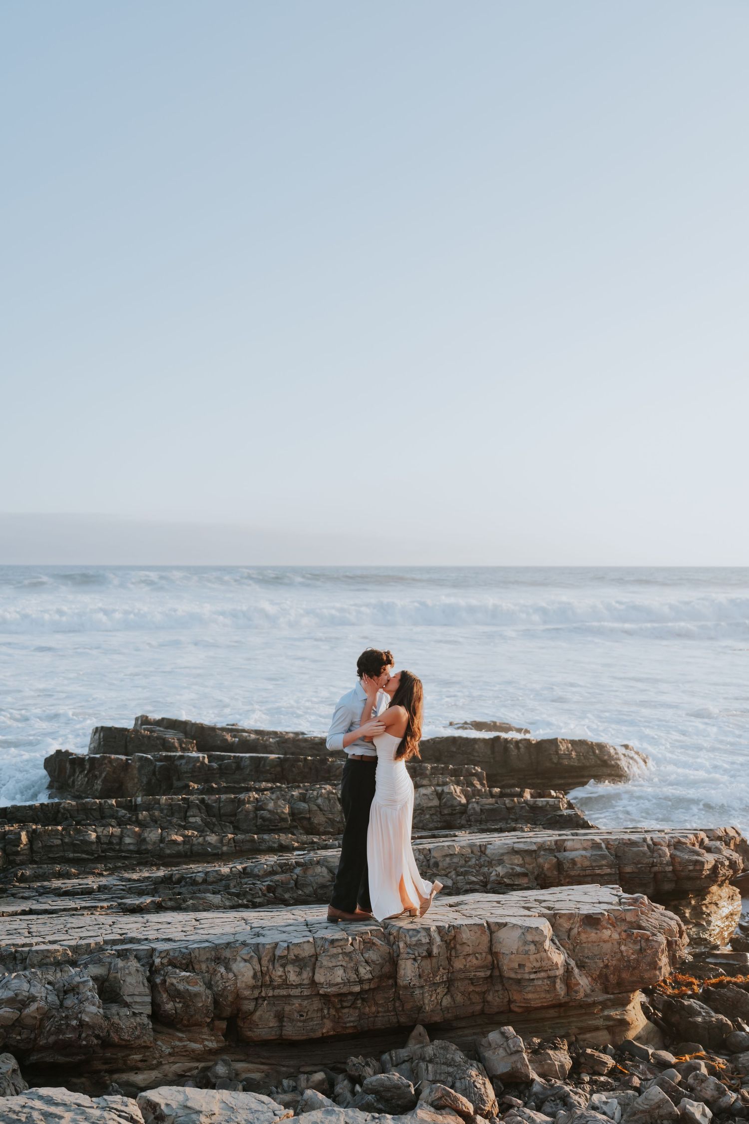 Couple embracing on rocky coastal cliffs with ocean waves crashing behind them at golden hour.