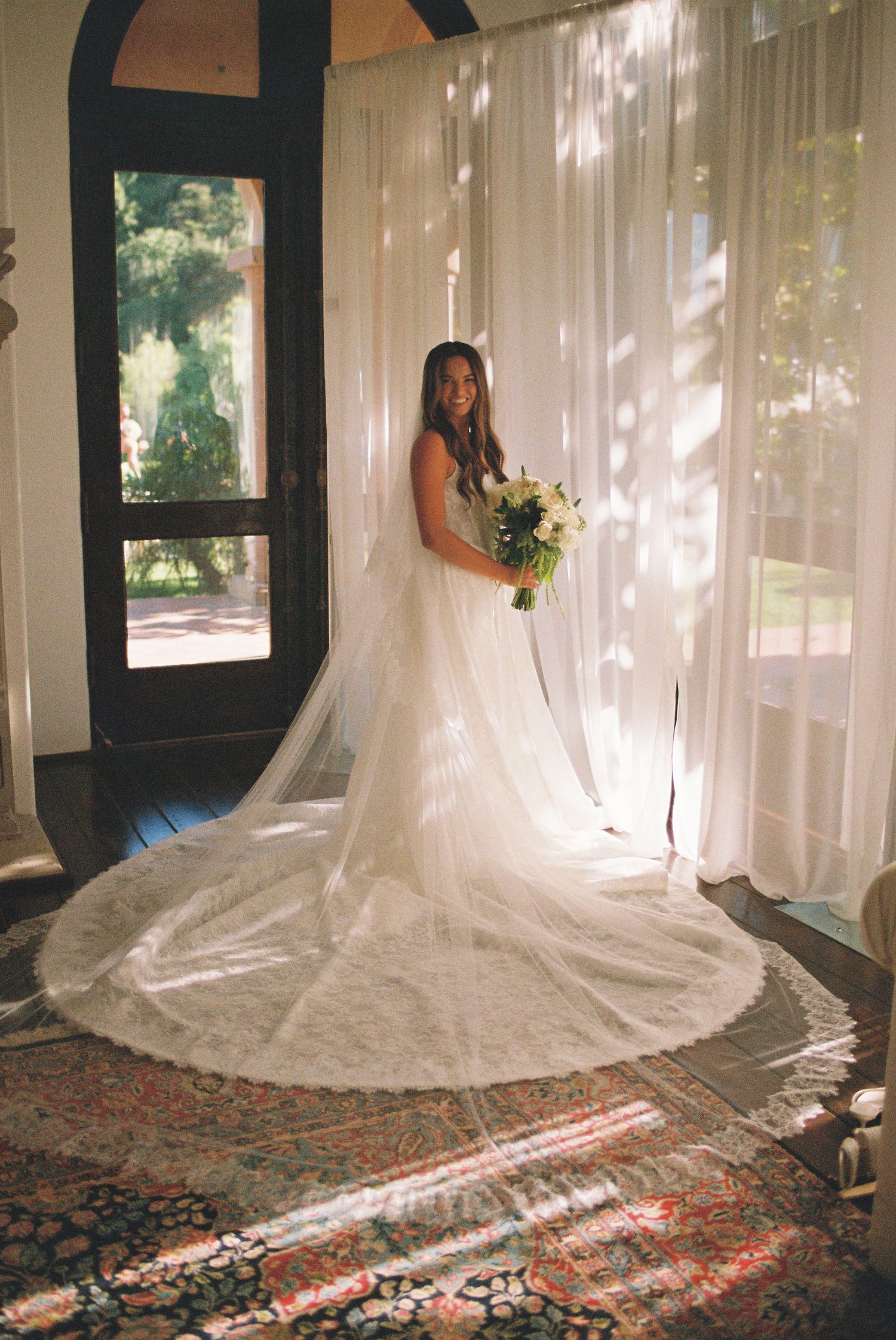 Bride in elegant lace wedding gown with long veil holds bouquet, standing before sheer white curtains in sunlit room.