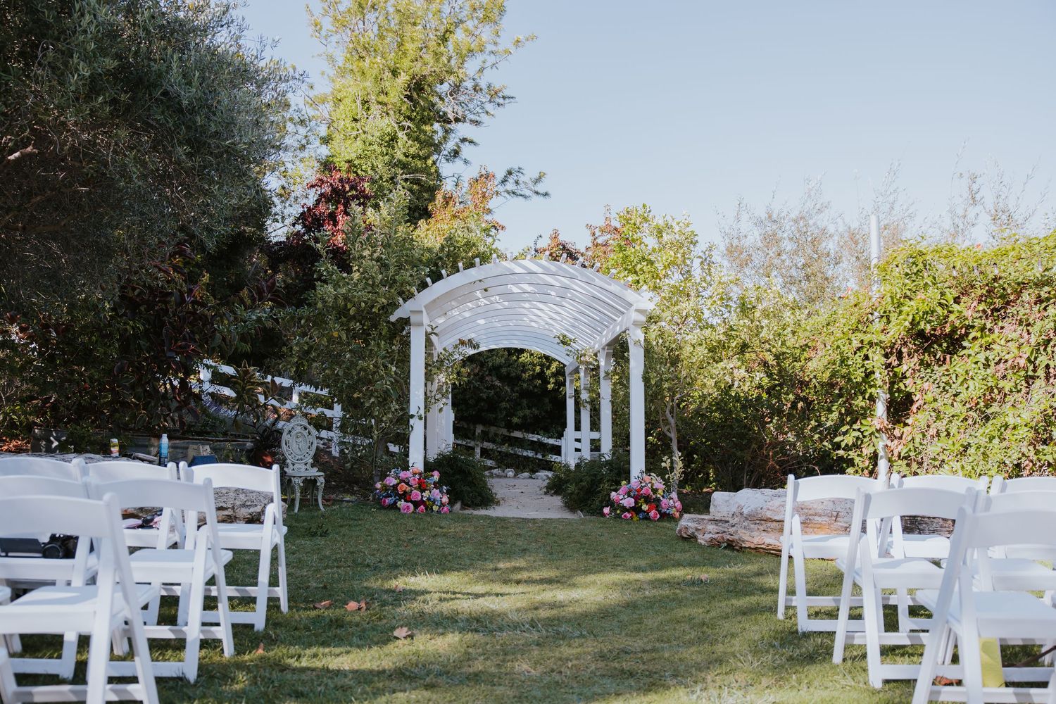White wedding ceremony setup with chairs and arch in an outdoor garden setting on a sunny day.