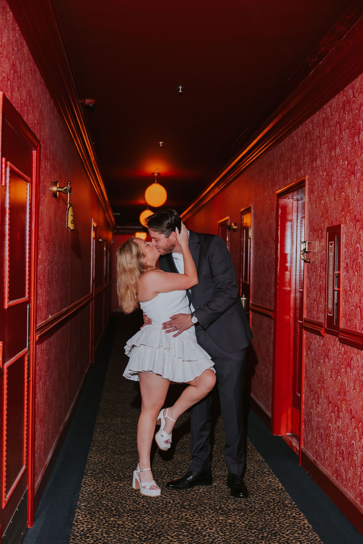 Newlyweds share a romantic kiss in a dramatic red wallpapered hallway with leopard print carpet.