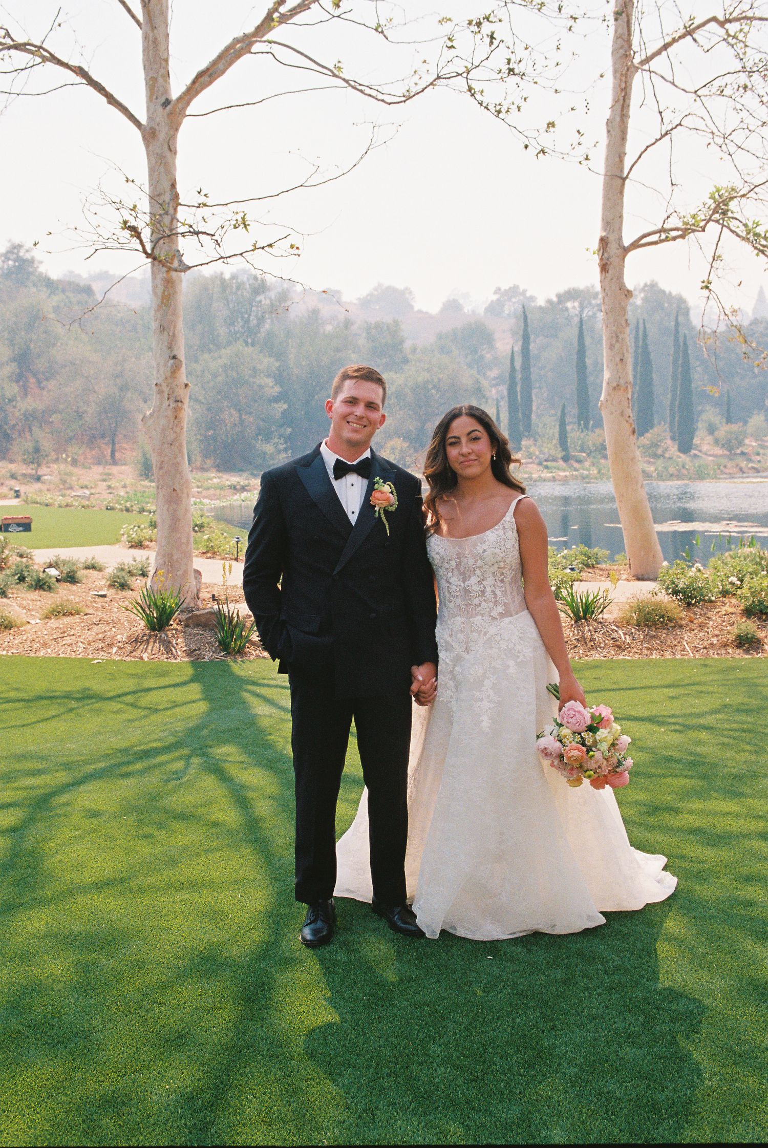 Bride and groom holding hands on green lawn, bride in lace gown with bouquet, groom in black tuxedo, trees and lake behind.