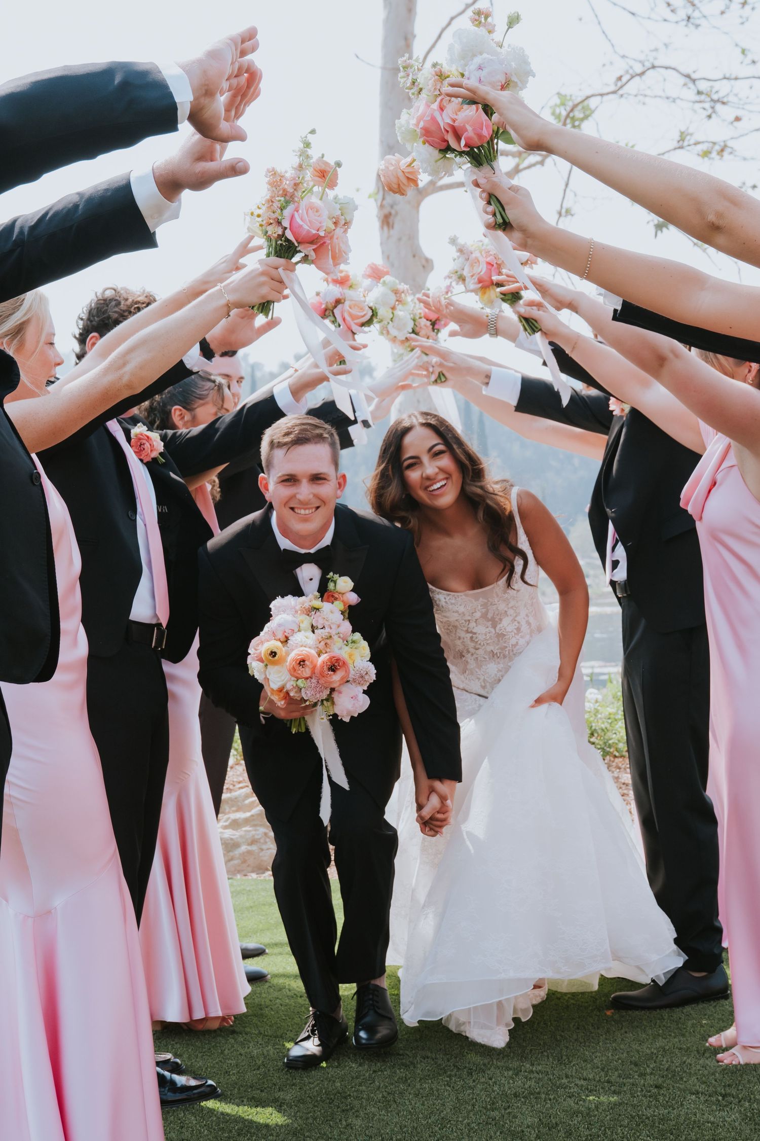 Bride and groom walk through floral arch tunnel with wedding party in pink dresses and black suits outdoors.