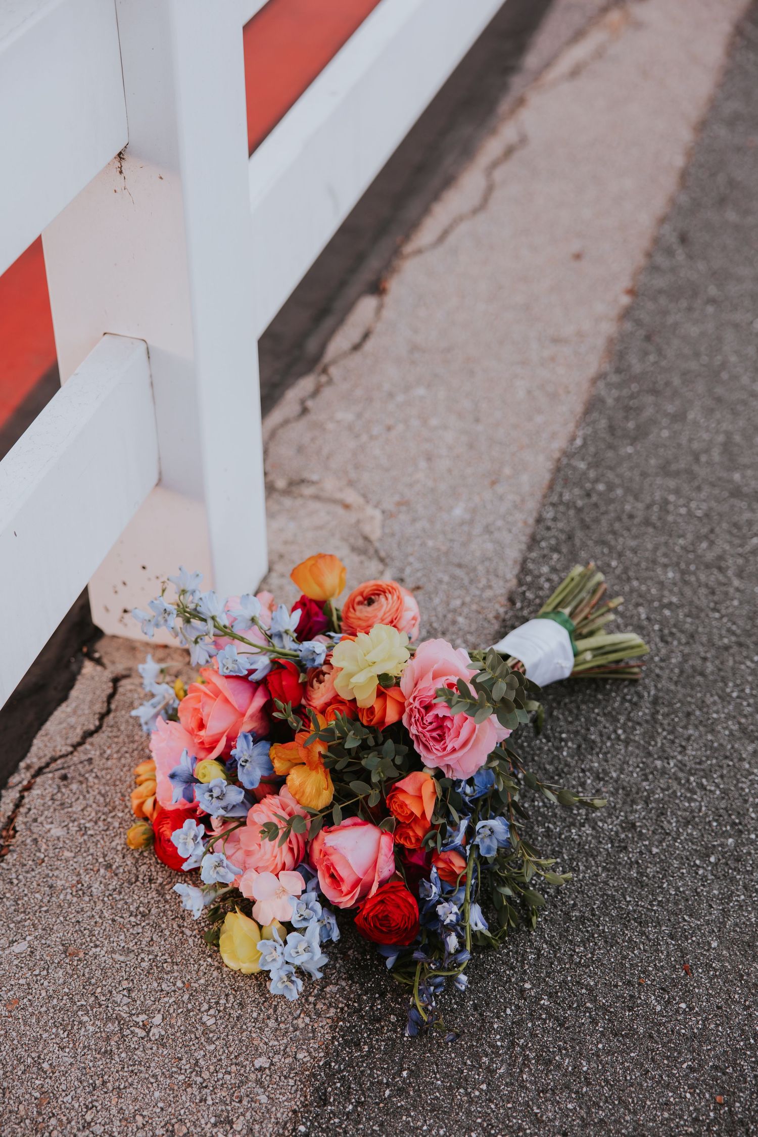 Vibrant floral bouquet with pink roses, blue hydrangeas, and orange flowers resting against a white fence.