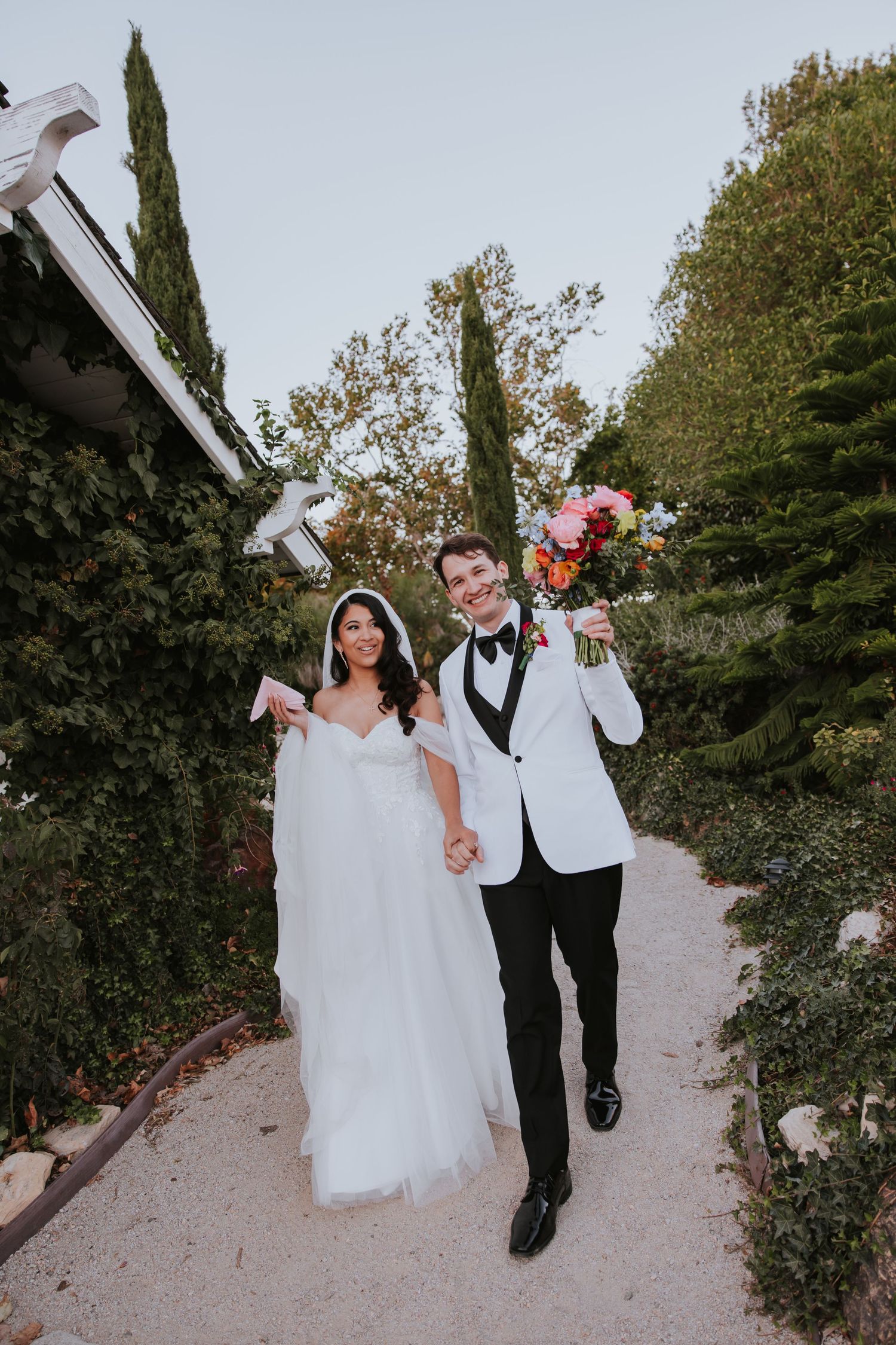 A joyful couple in wedding attire playfully walks down a path surrounded by greenery, holding hands and laughing together.