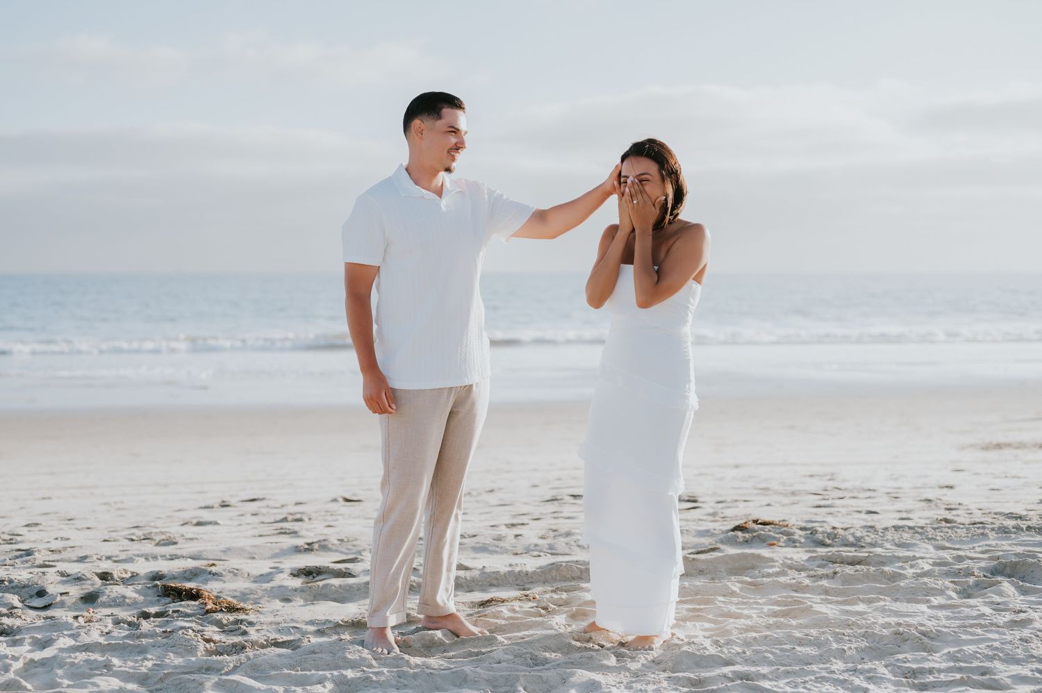 A couple in white outfits walks together on a sandy beach during sunset, creating romantic silhouettes against the ocean.