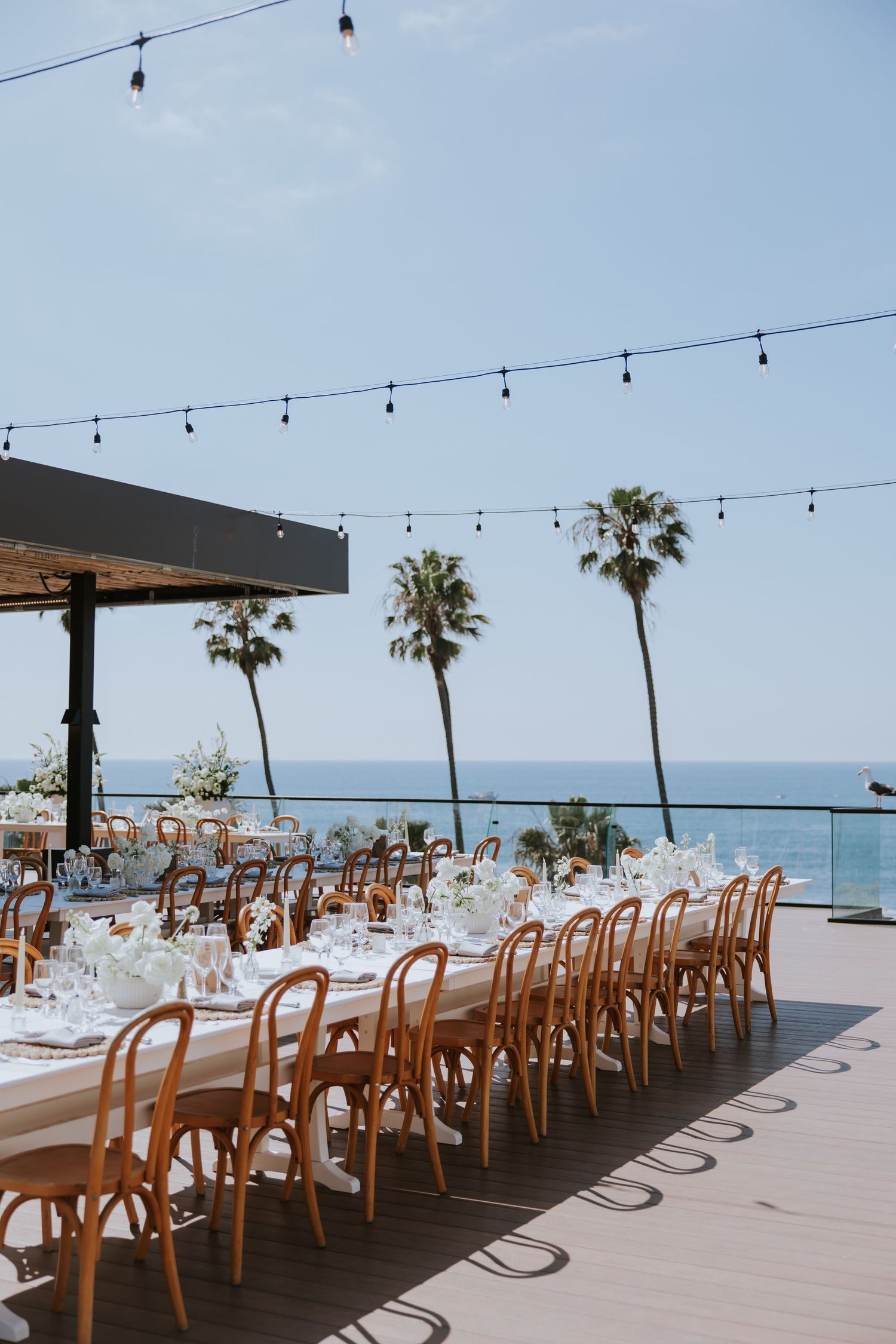 Outdoor wedding reception setup with wooden chairs and string lights overlooking the ocean.