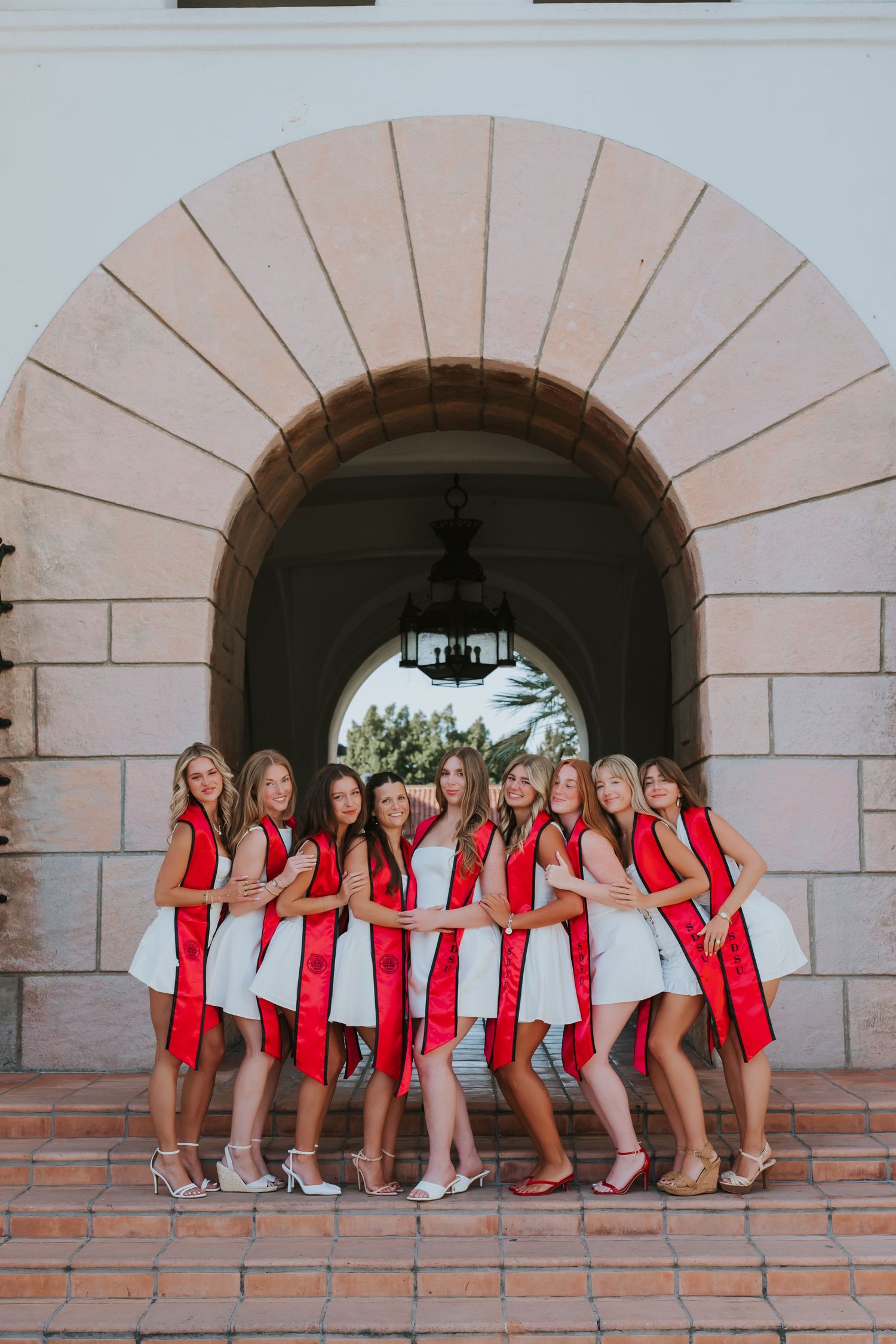 Group of young women in matching white dresses and red sashes posing under a stone arch for a sorority photo.