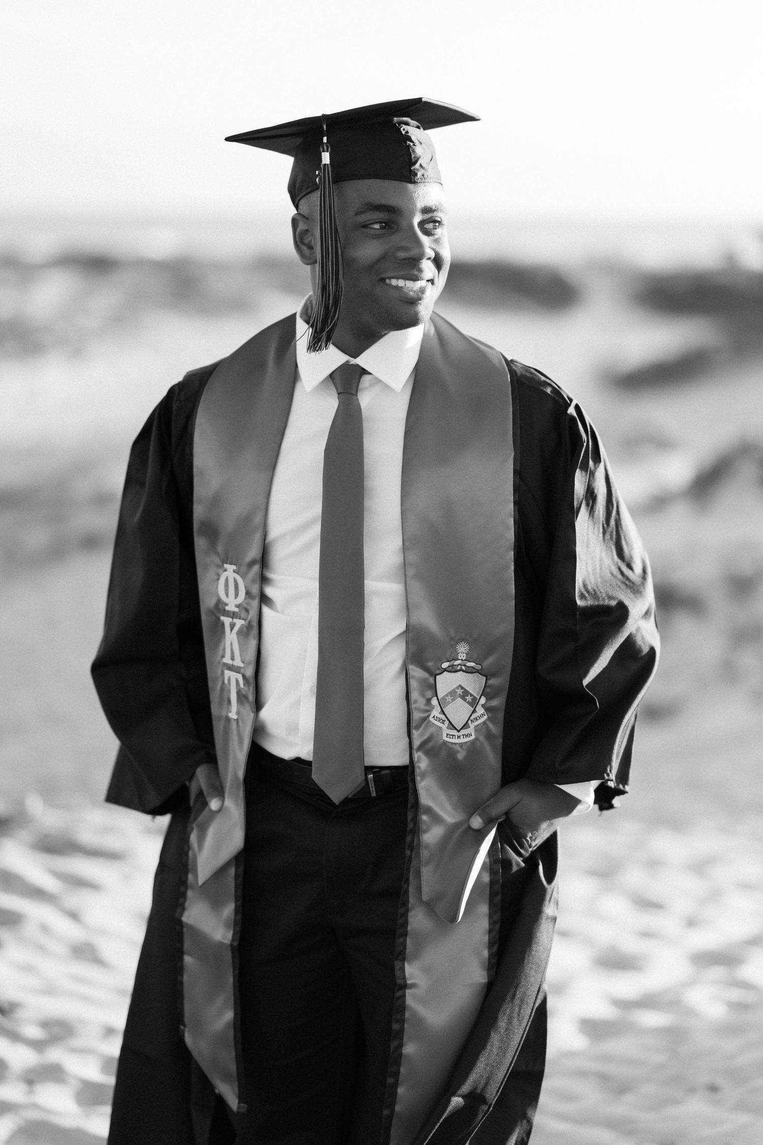 Young man in graduation cap and gown with Phi Kappa stole smiling outdoors in black and white photo.