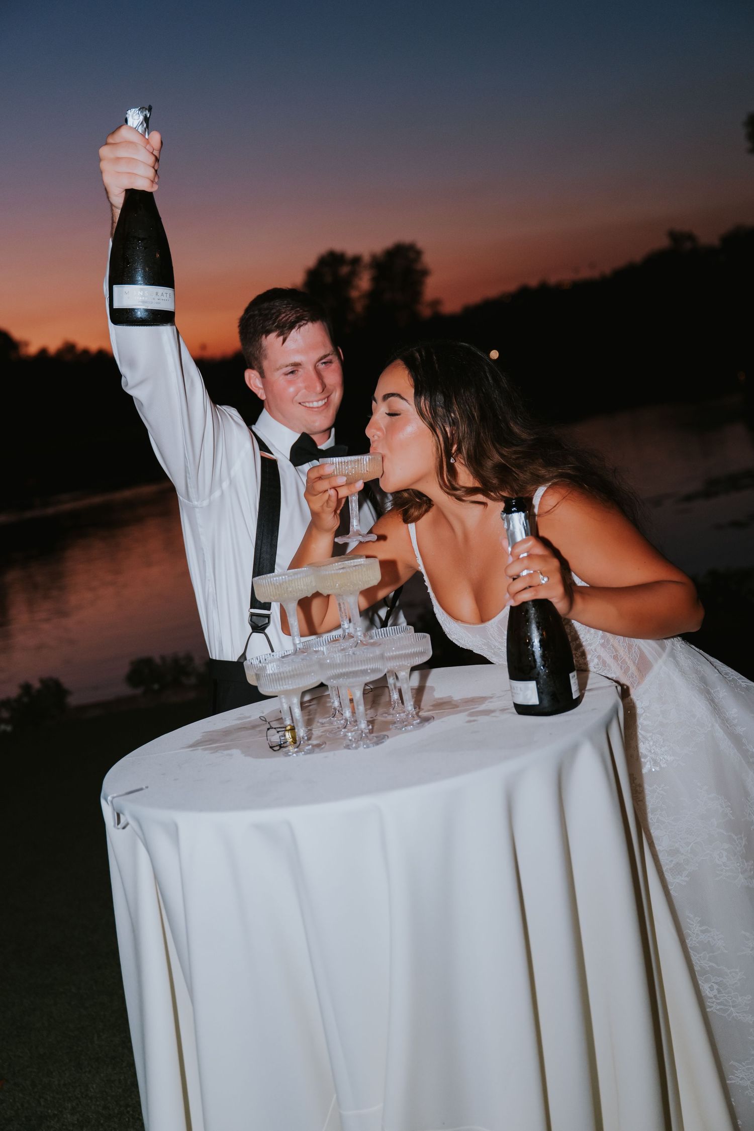 Bride and groom celebrating at sunset, popping champagne bottles at a tower of glasses outdoors near a lake.
