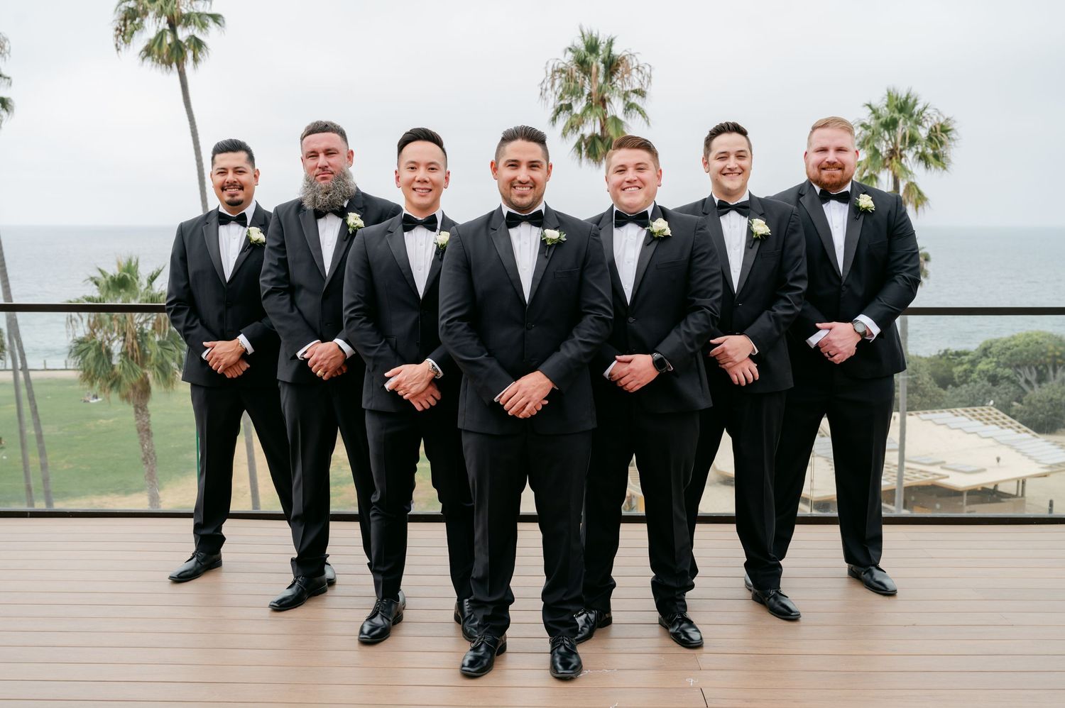 Groomsmen in black tuxedos and bowties pose together on oceanfront deck.