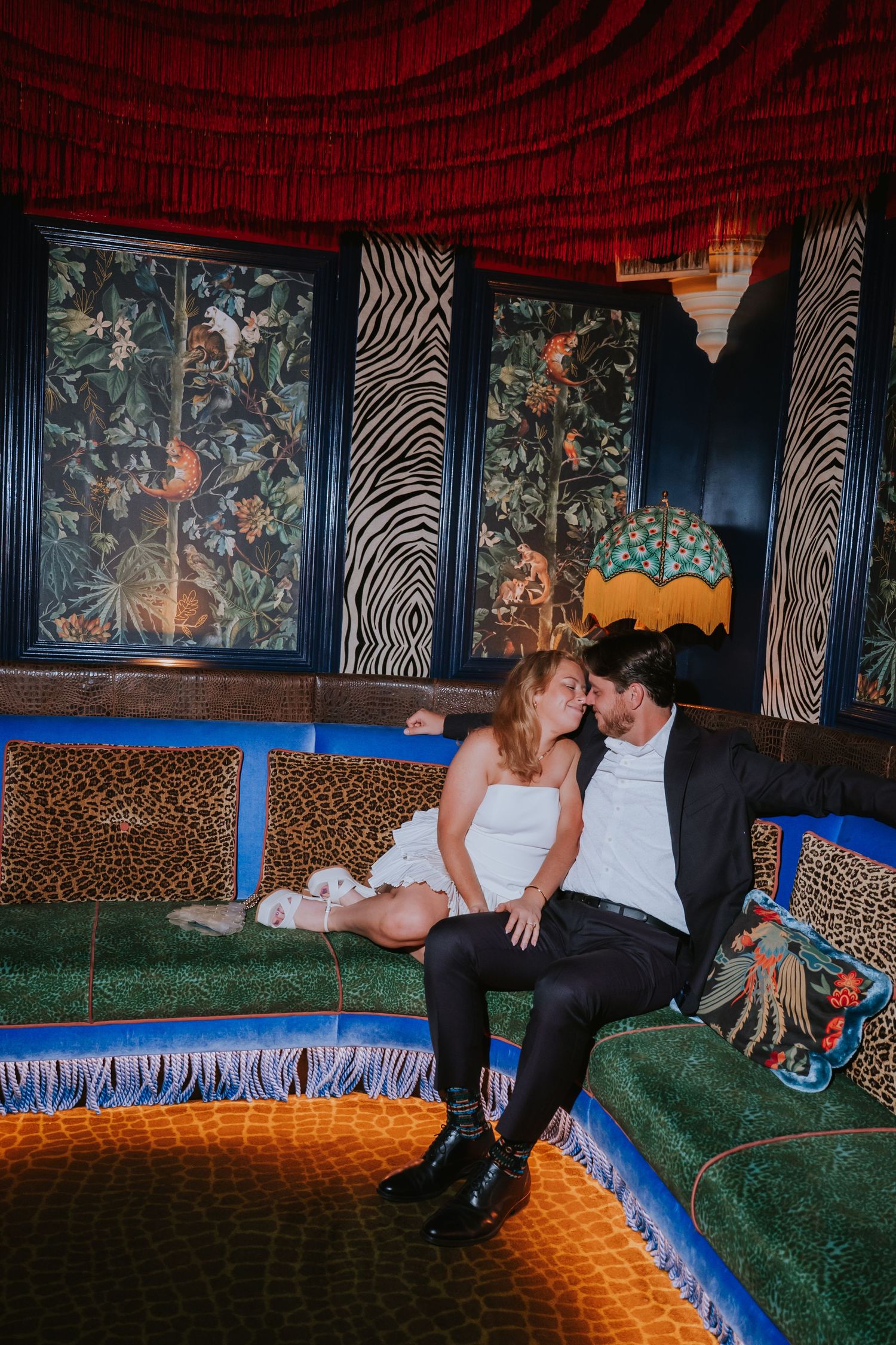 Couple sits intimately on a colorful eclectic sofa surrounded by zebra print and jungle wallpaper.
