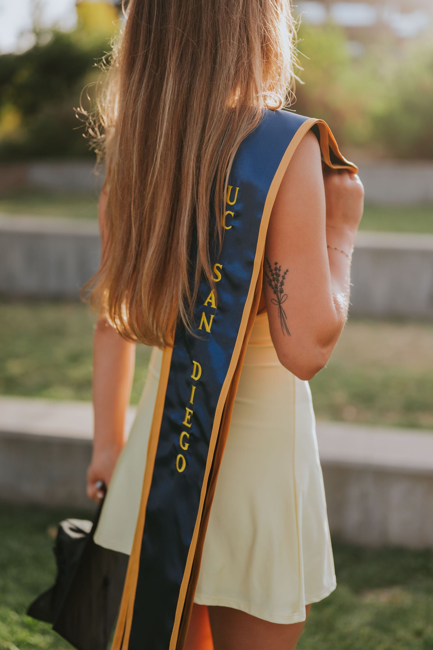 UC San Diego graduate wearing navy and gold graduation sash over white dress, showing tattoo on arm.