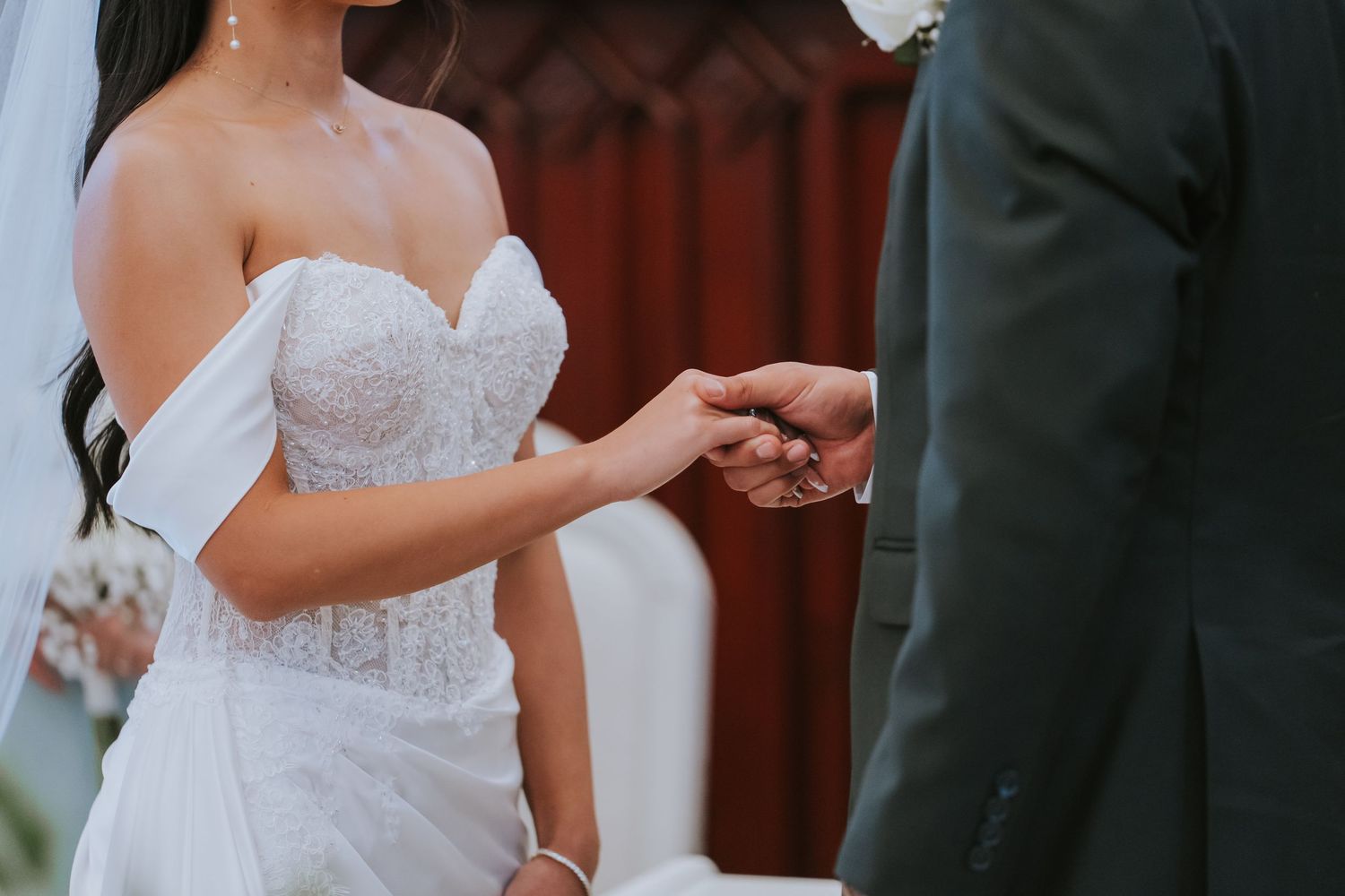 Close up of hands exchanging rings during outdoor wedding ceremony with white lace bridal gown visible.