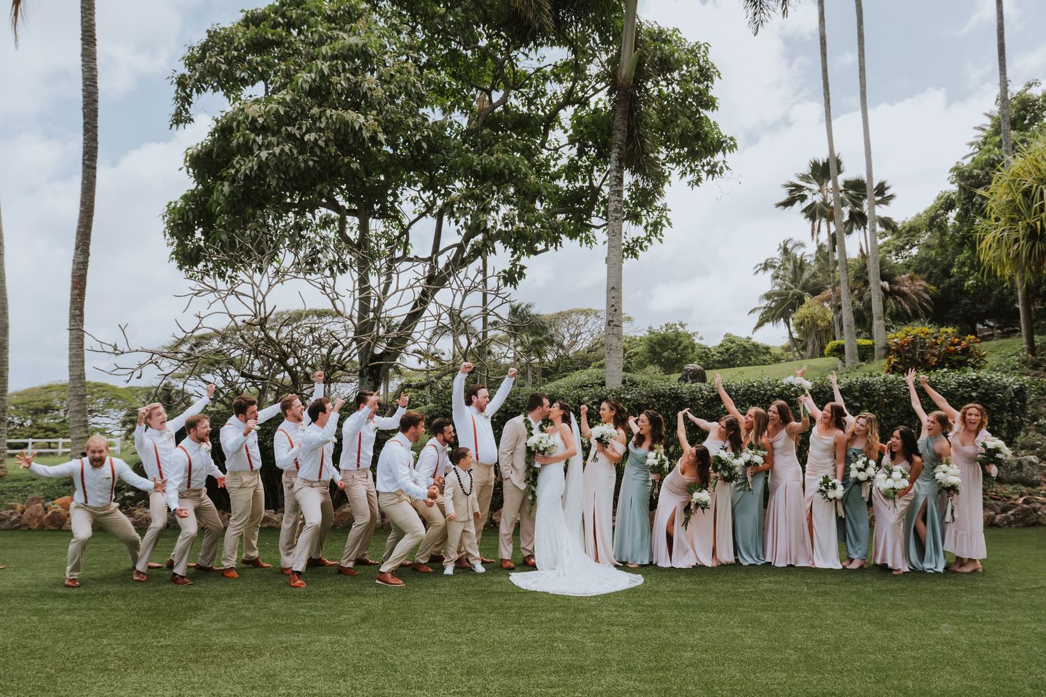Wedding party in formal attire walking together on a grassy lawn with palm trees in the background.