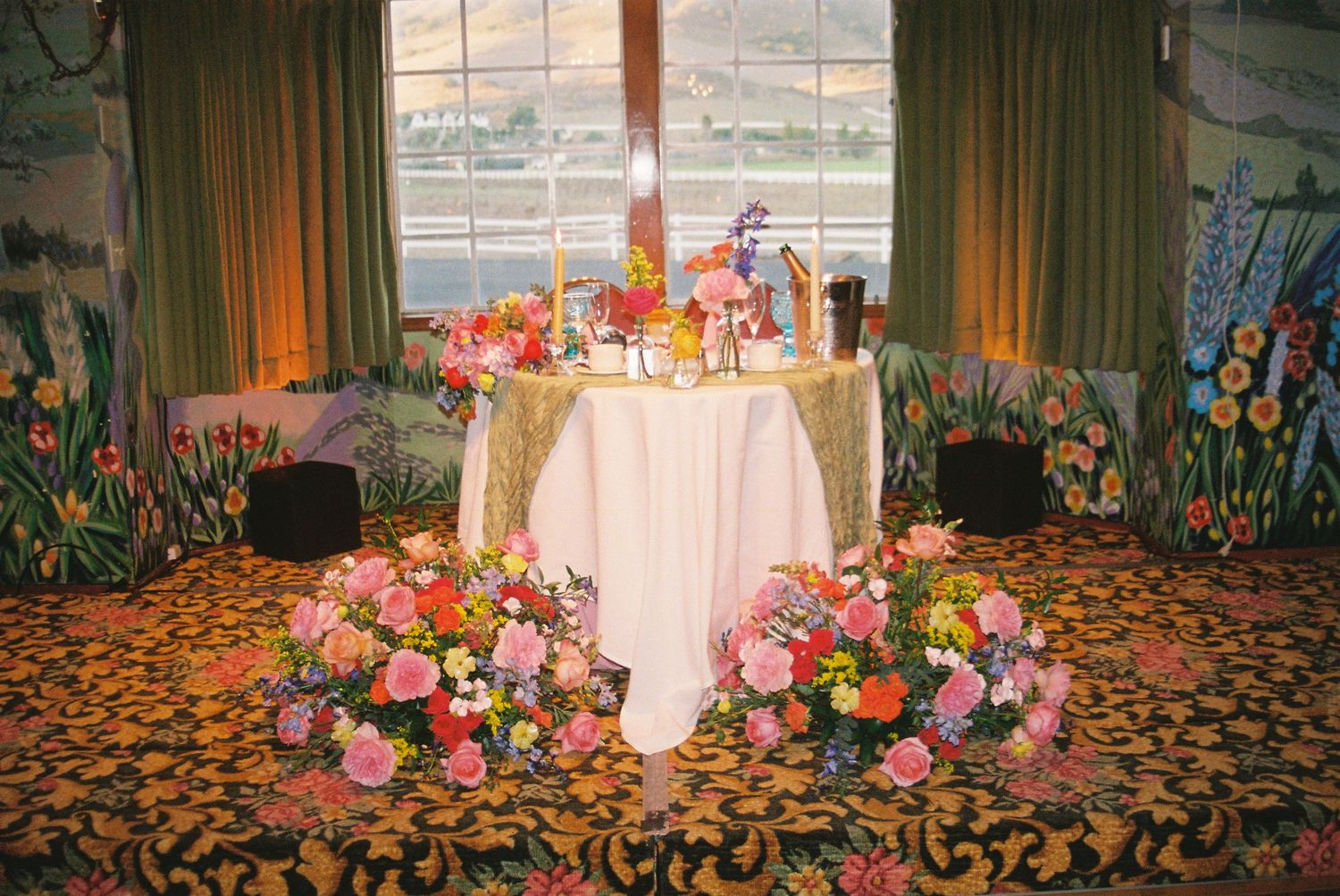 Colorful floral arrangements adorn round tables in an elegant ballroom with patterned carpet and green drapes.