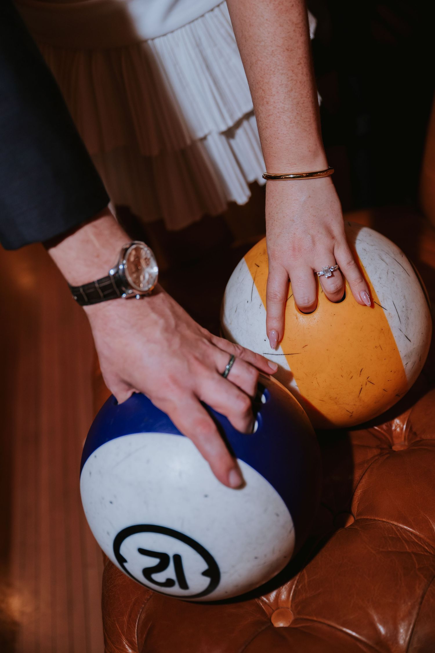 Close-up of newlyweds hands with wedding rings resting on oversized bowling balls at a wedding reception.