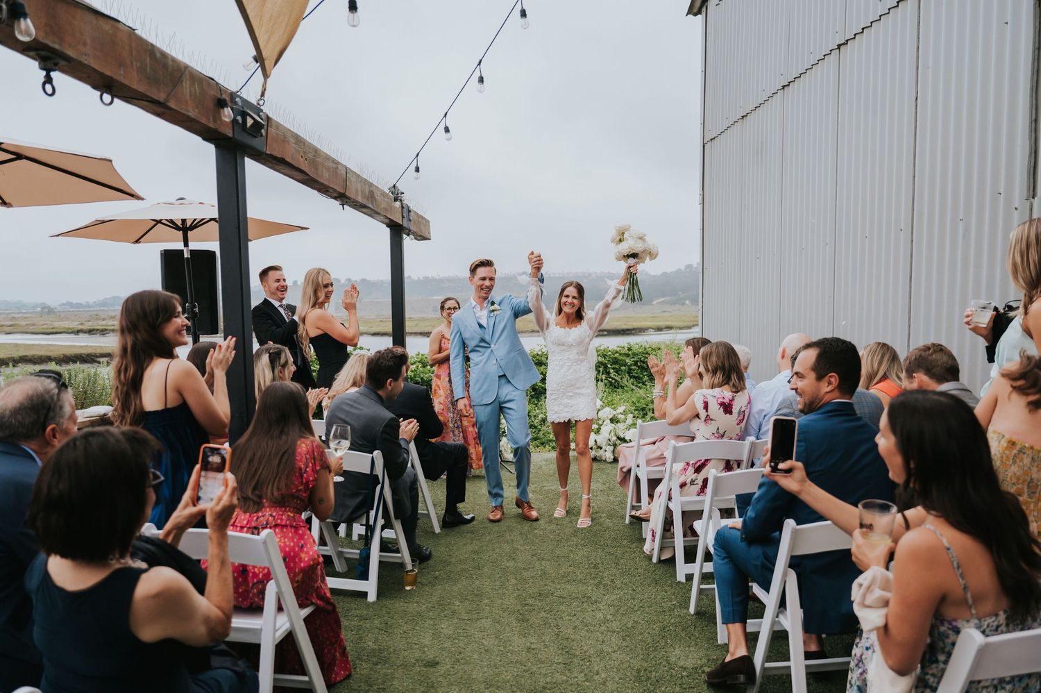 Outdoor wedding reception setup with white chairs and string lights at a rustic barn venue.