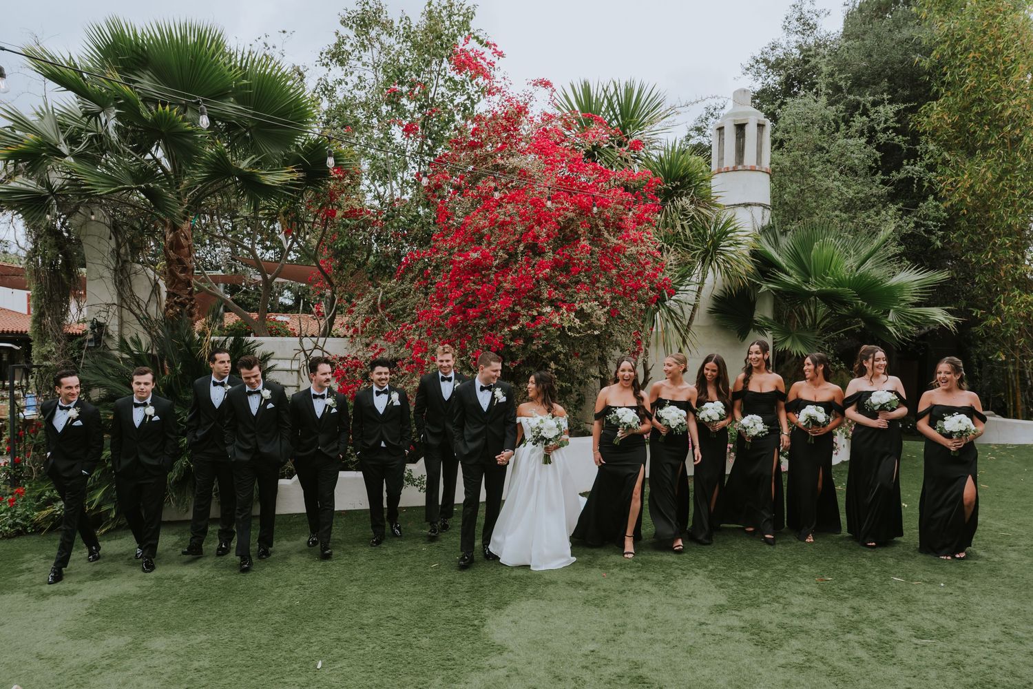 Wedding party members lined up outdoors against vibrant red bougainvillea flowers.