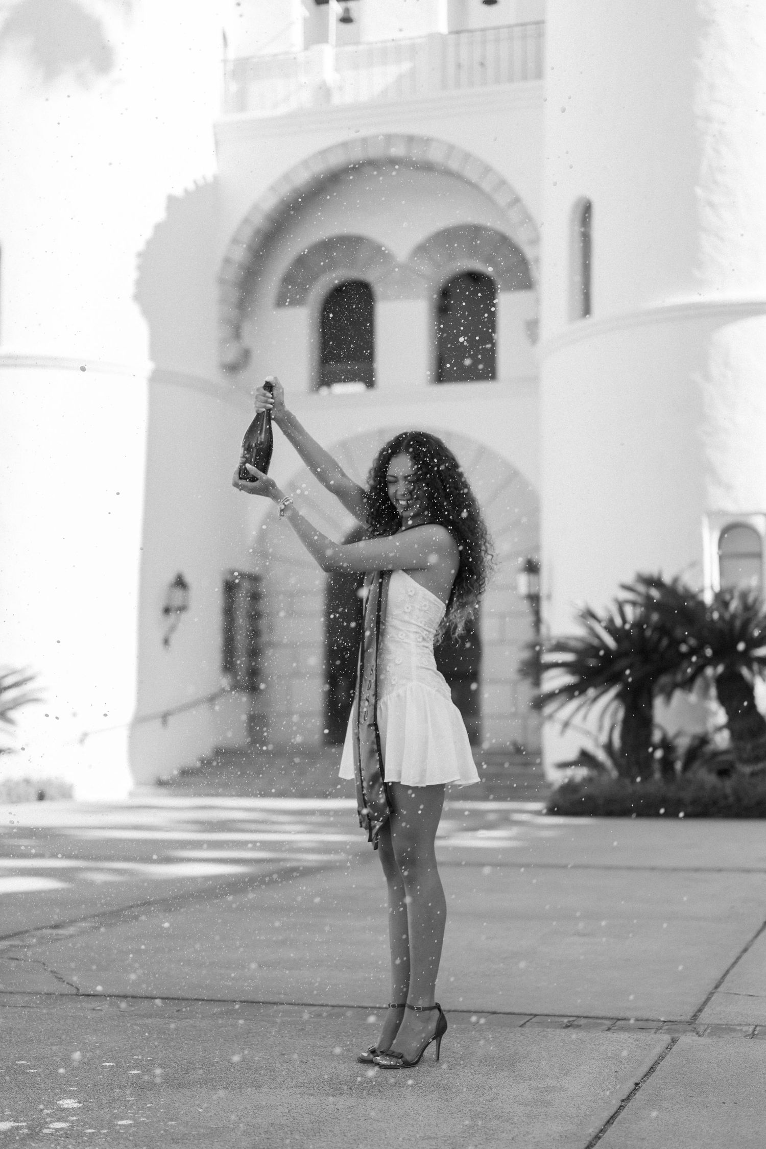 A woman in a white dress poses gracefully in front of a white Mediterranean-style building with arches.