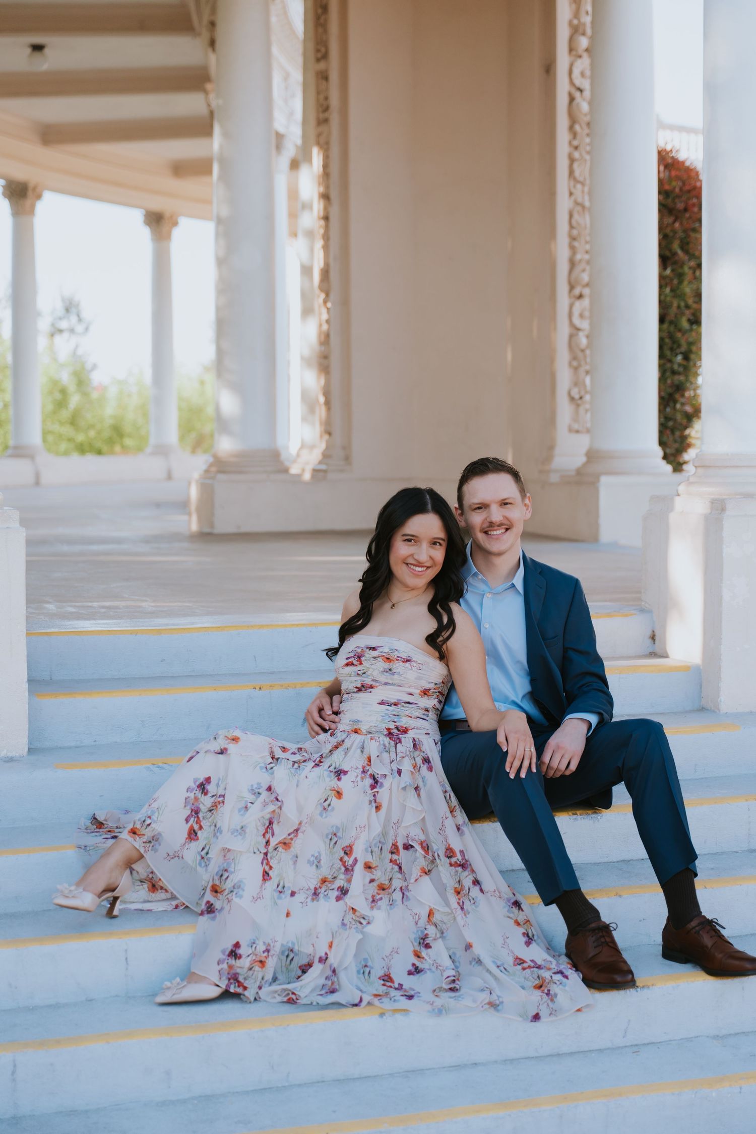 Couple sitting on elegant white marble steps, woman in floral dress and man in navy suit, posing at a classical columned building.