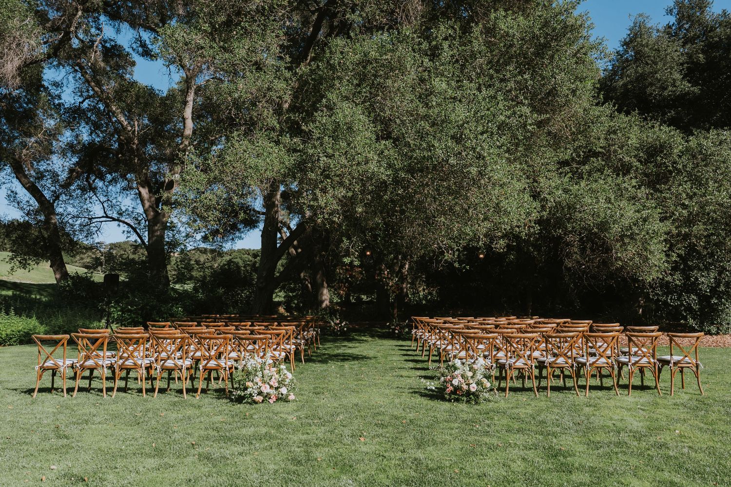 Outdoor wedding ceremony setup with wooden chairs on green lawn surrounded by lush trees and floral arrangements.