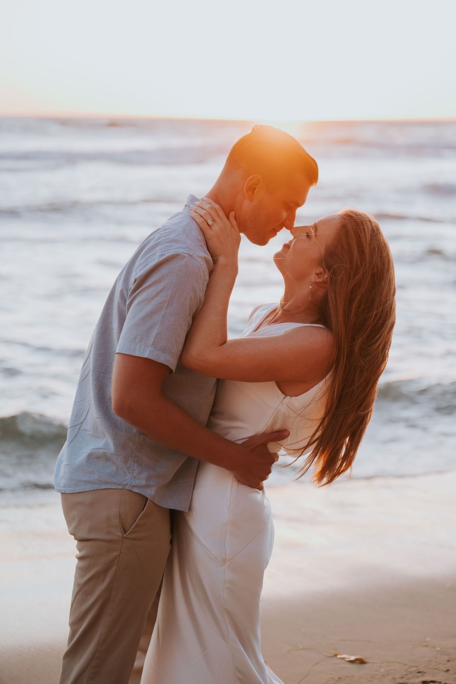 Couple sharing a romantic sunset kiss on the beach, waves crashing behind them in golden light.