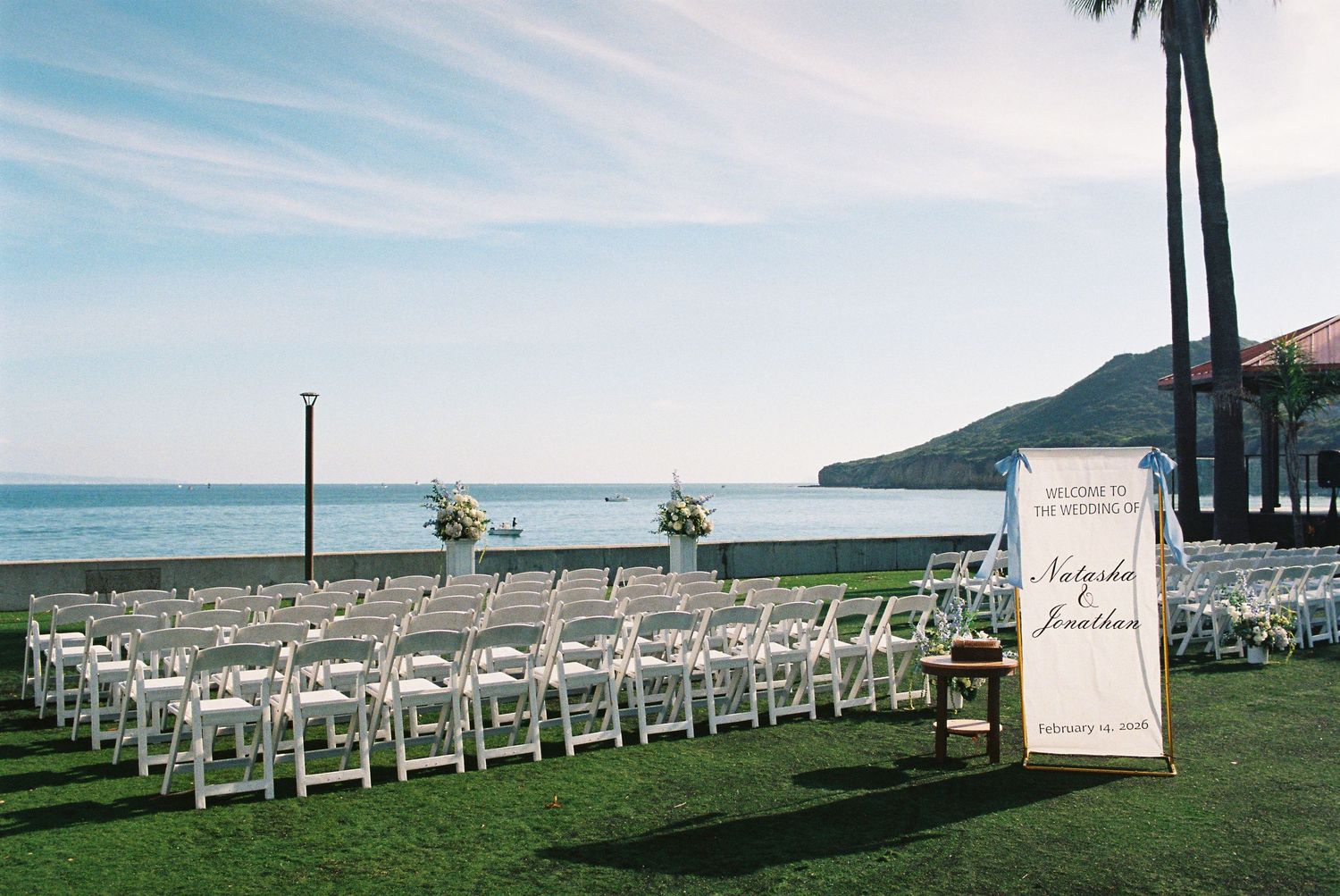 Outdoor wedding ceremony setup with white chairs on green lawn overlooking ocean, palm trees, and mountains under blue sky.