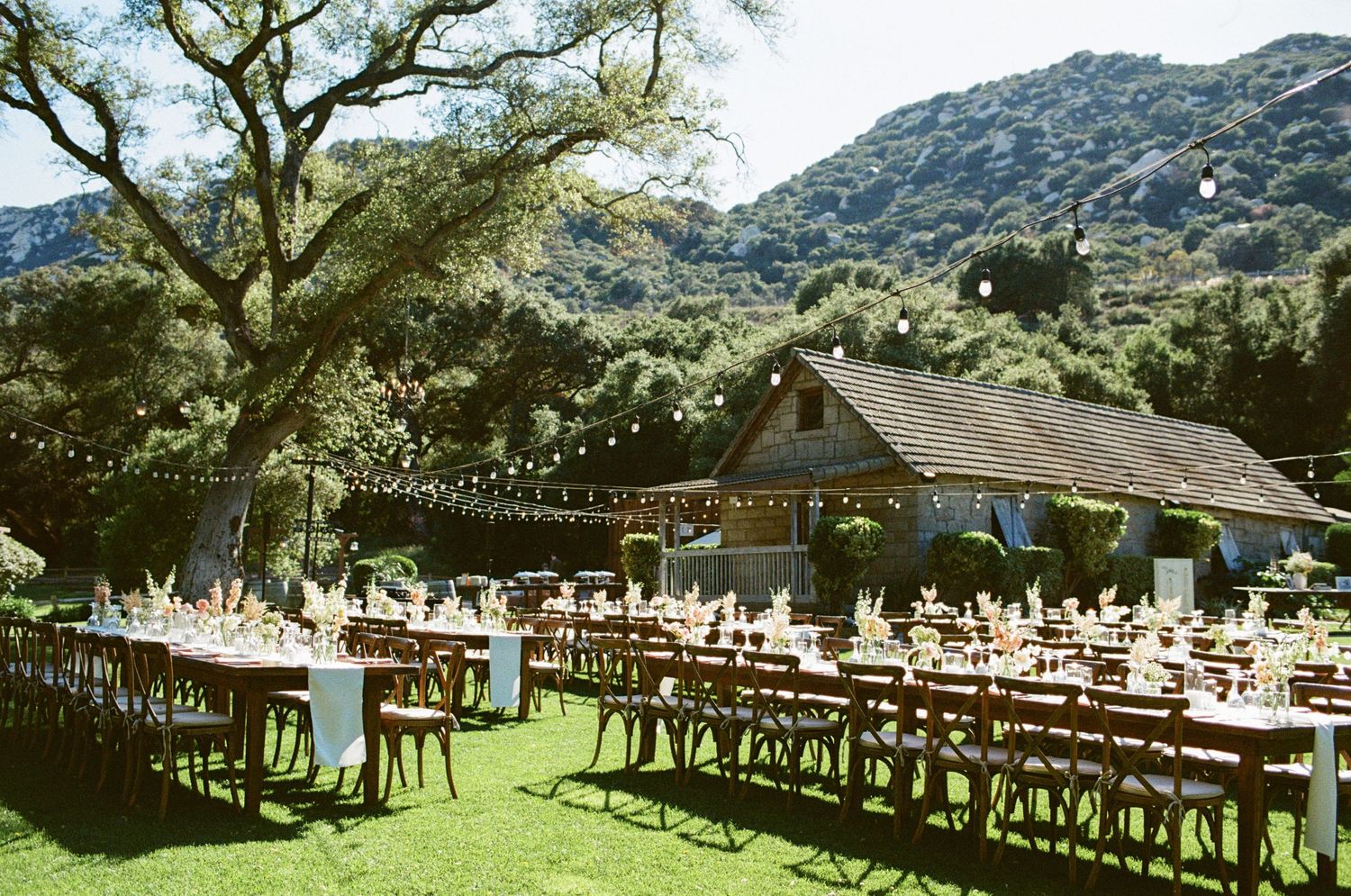 Elegant outdoor wedding reception setup with string lights, long tables, and mountain backdrop.