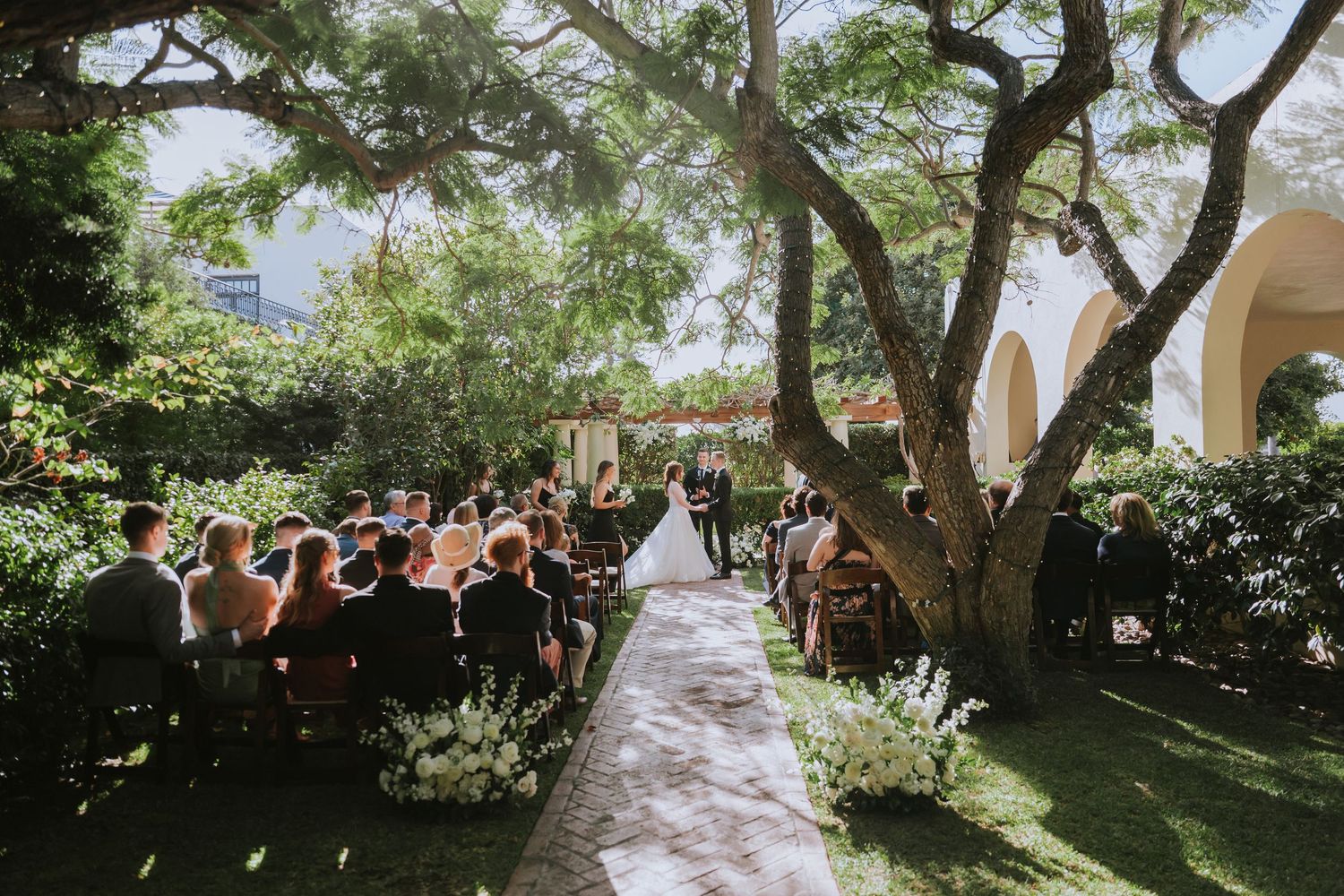 String lights illuminate an outdoor garden wedding ceremony path lined with trees and chairs.
