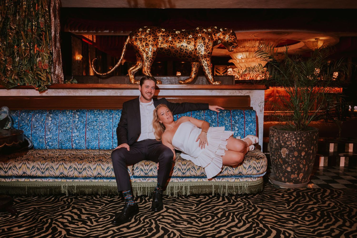 Couple relaxes on a patterned sofa beneath a golden leopard statue in a luxurious venue.