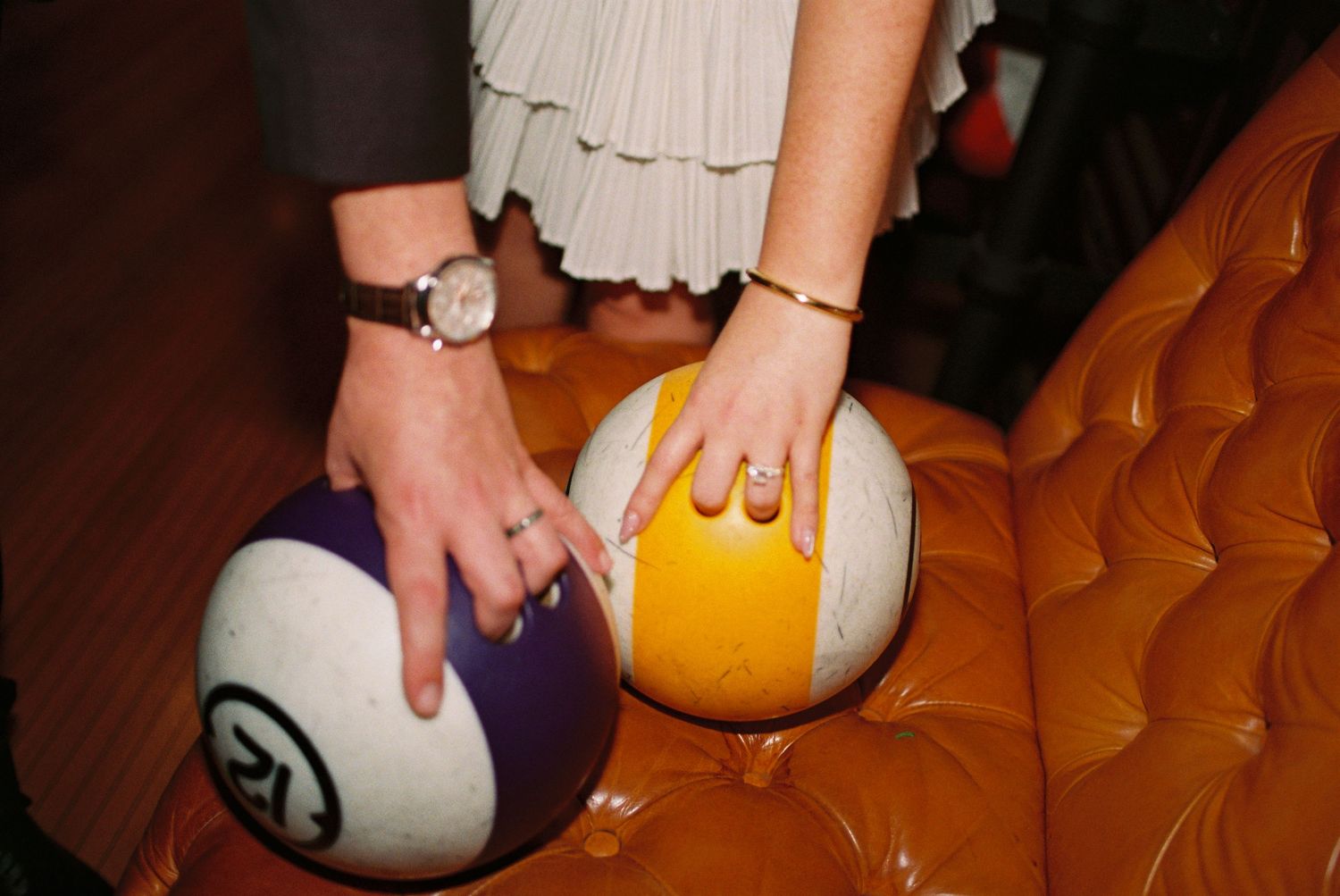 Two hands resting on colorful billiard balls on an orange tufted leather couch, wearing rings and bracelets.