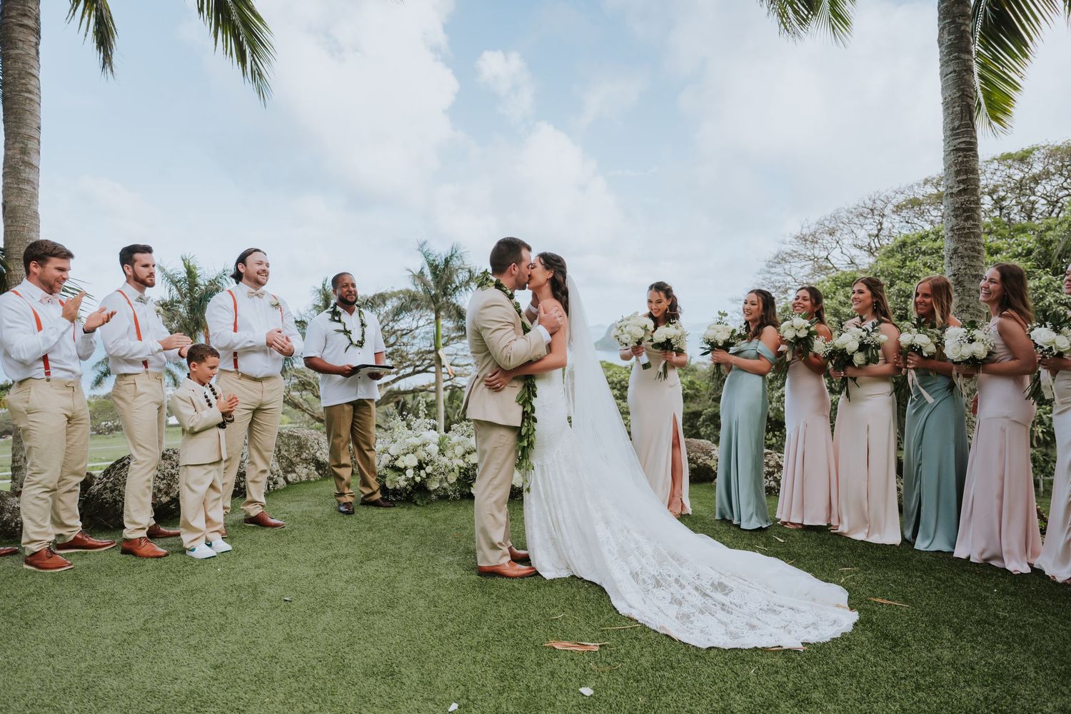 Wedding party gathers on a tropical lawn with palm trees as couple shares their first kiss as newlyweds.