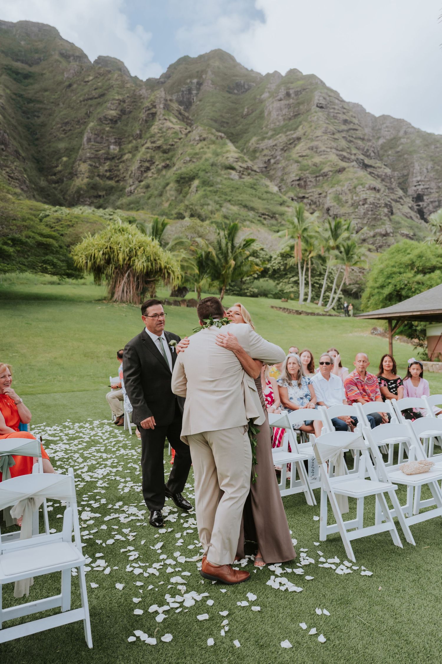 Wedding ceremony setup with scattered flower petals and guests seated on white chairs against mountain backdrop.